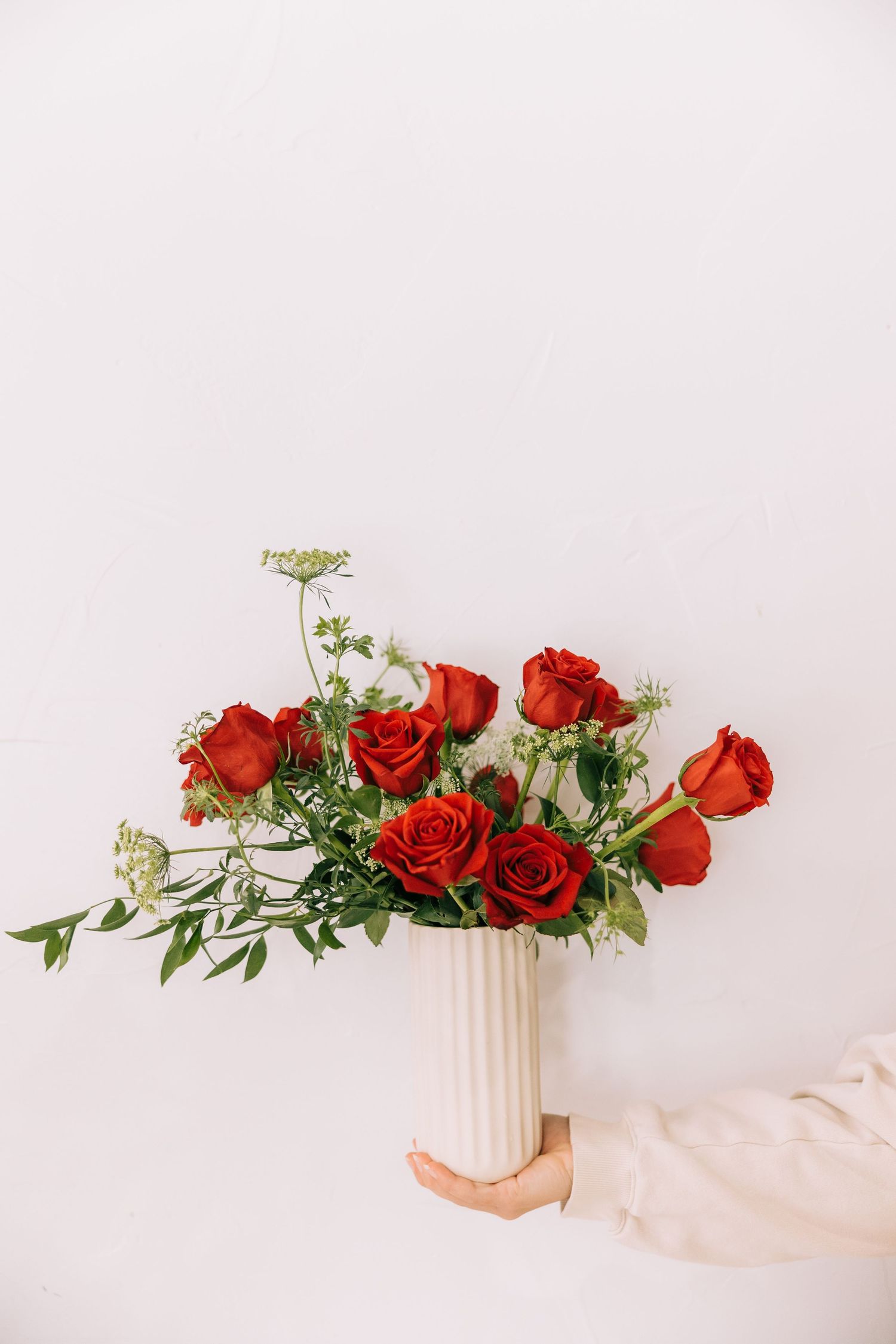 Hand holding a white ribbed vase with vibrant red roses and delicate greenery against a light background.