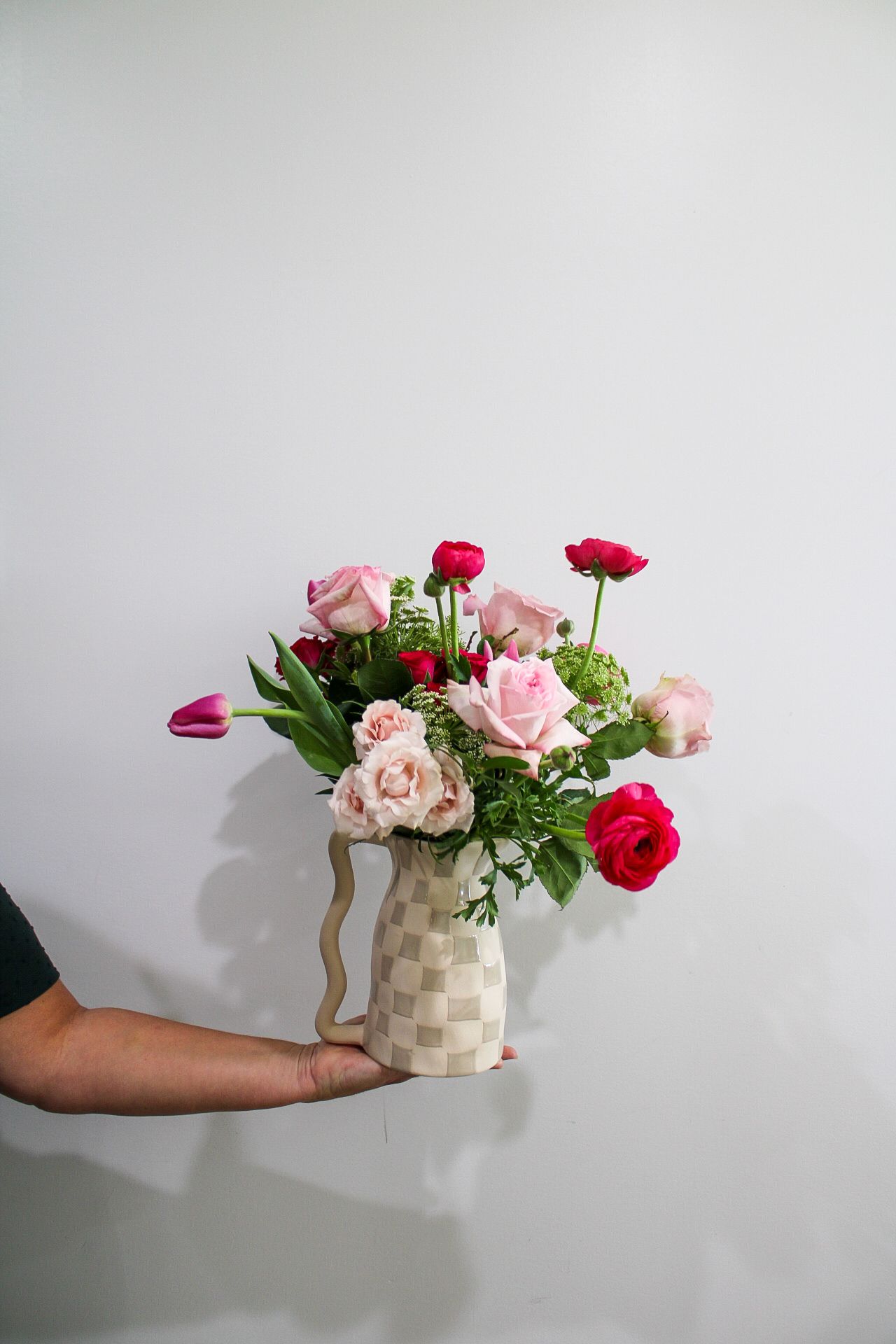 Hand holding a checkered vase with pink and red roses, ranunculus, and tulips against a white wall.