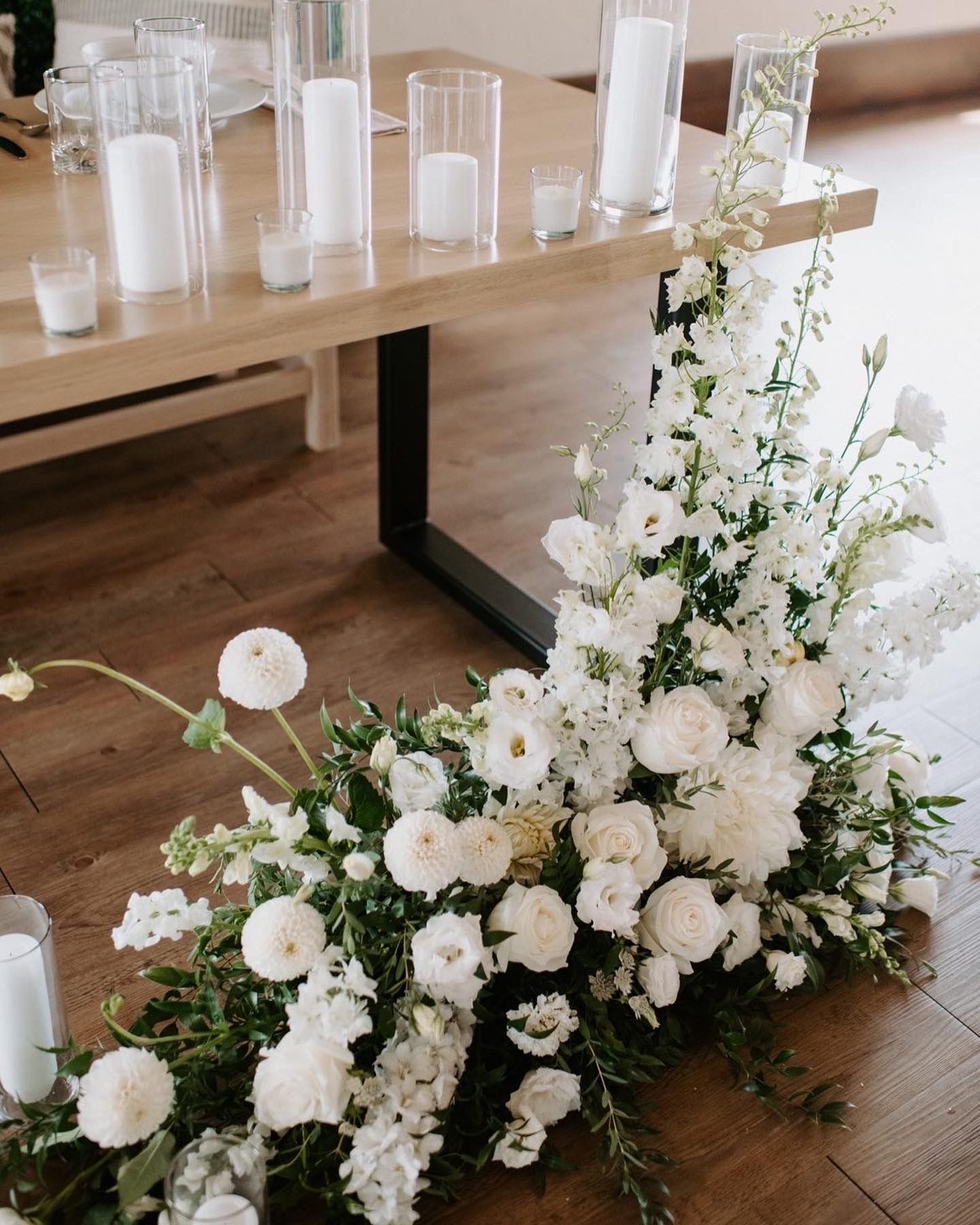 Luxurious white floral arrangement cascading down wooden table with glass candle holders.
