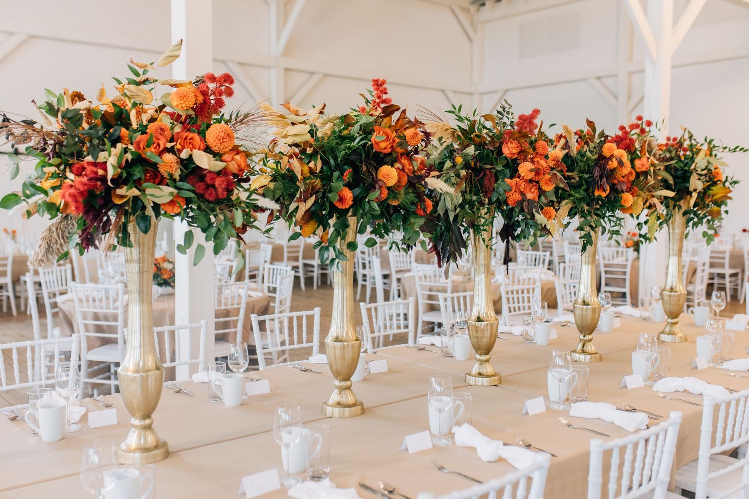 Elegant wedding reception table setup with tall gold centerpieces featuring vibrant orange and red floral arrangements.