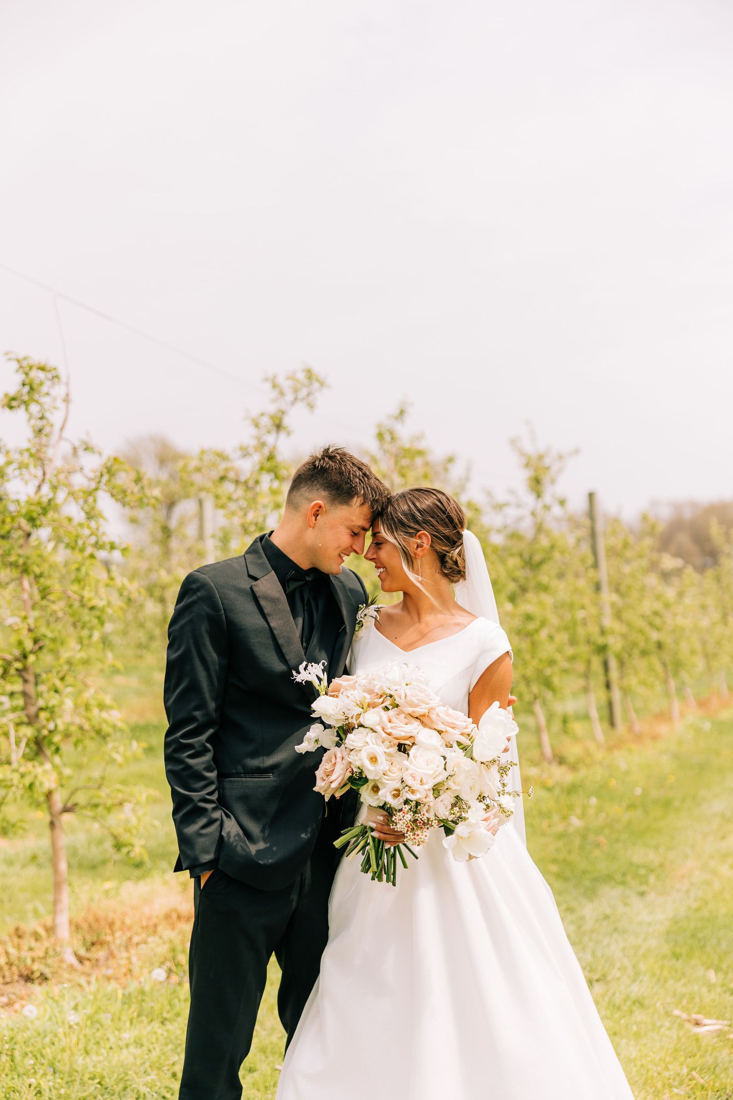 Elegant wedding couple shares intimate moment in orchard with white floral bouquet.
