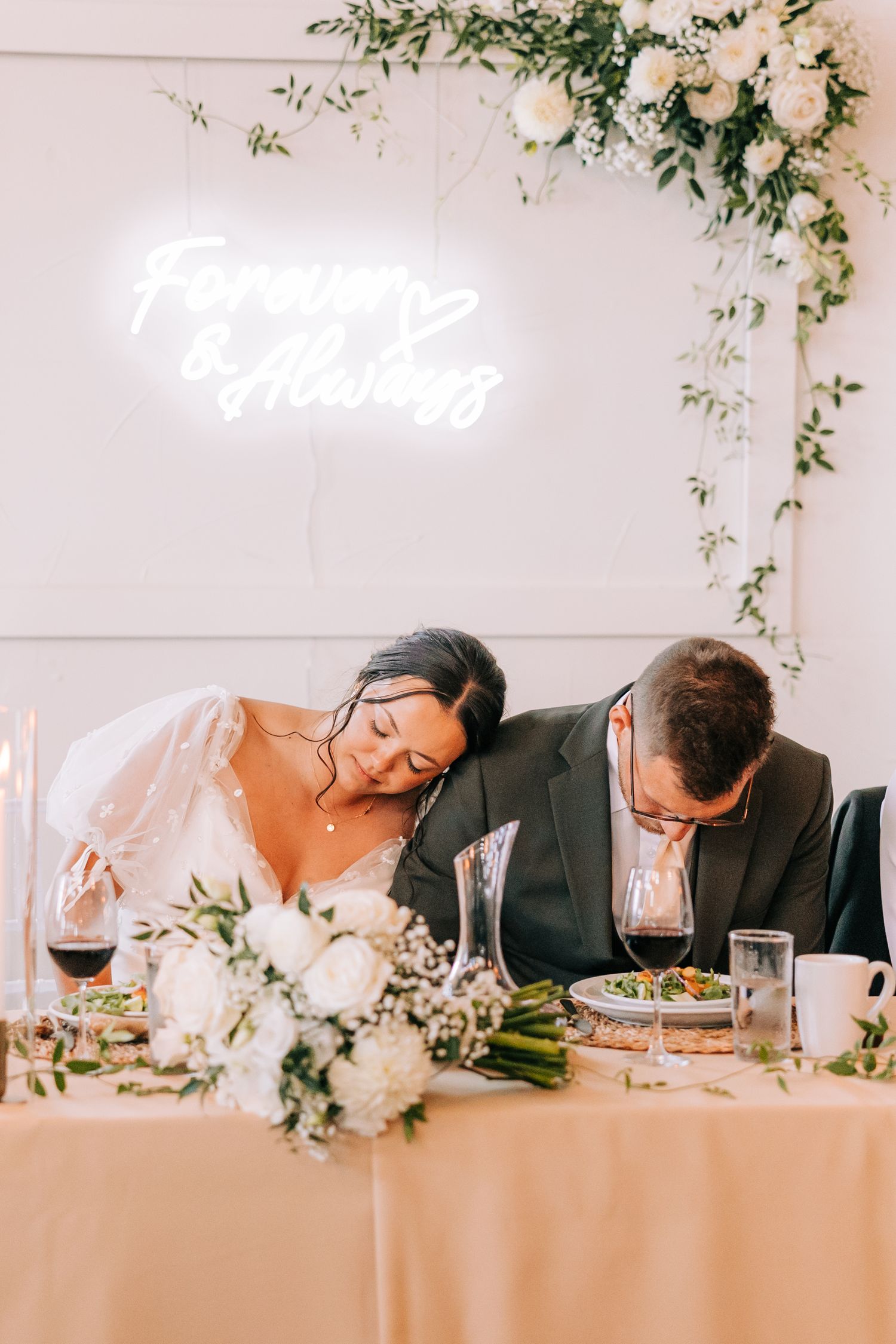 Reception table with neon sign, white floral arrangements and wine glasses.