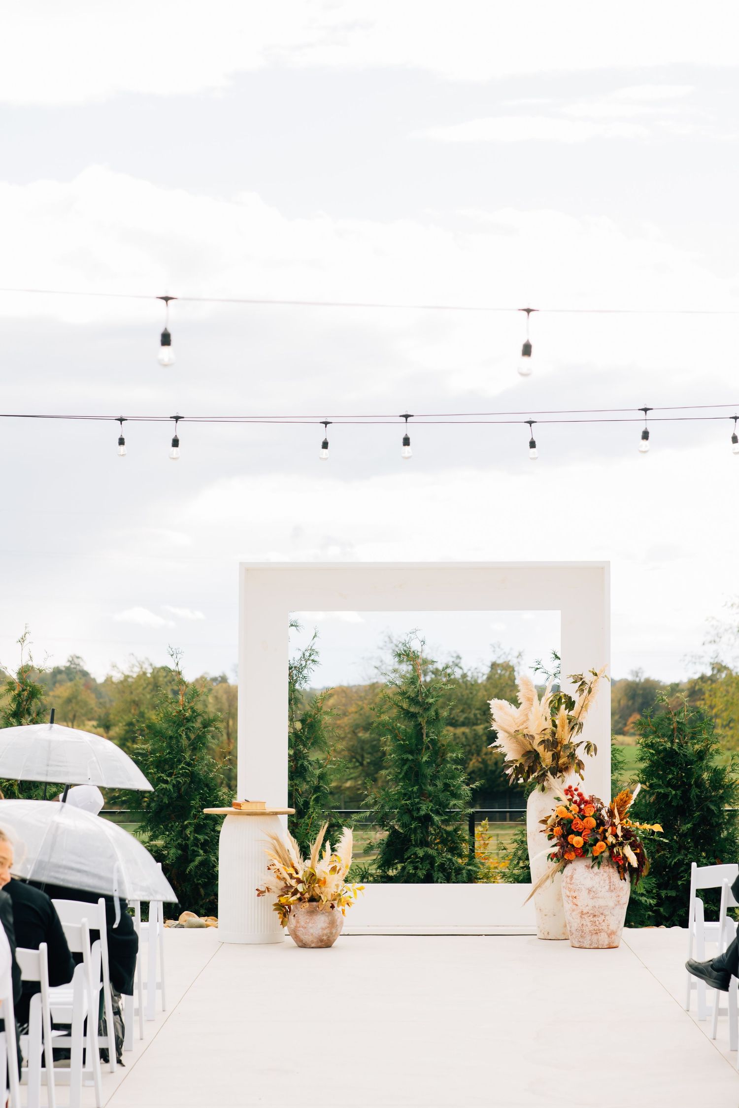 Modern white wedding arch with autumn floral arrangements and string lights overhead.