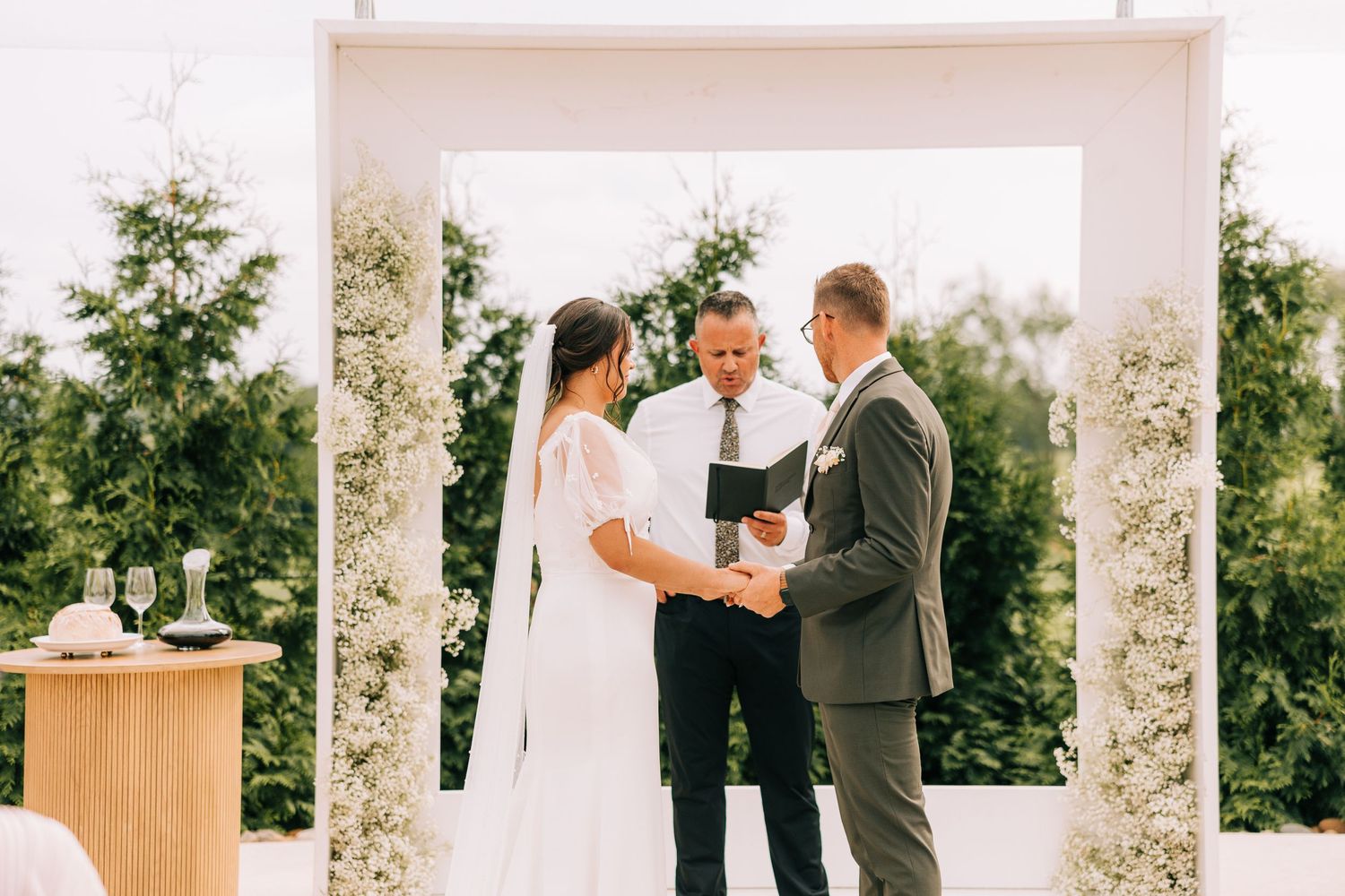 An elegant outdoor wedding ceremony under a white floral arch with pastoral views.