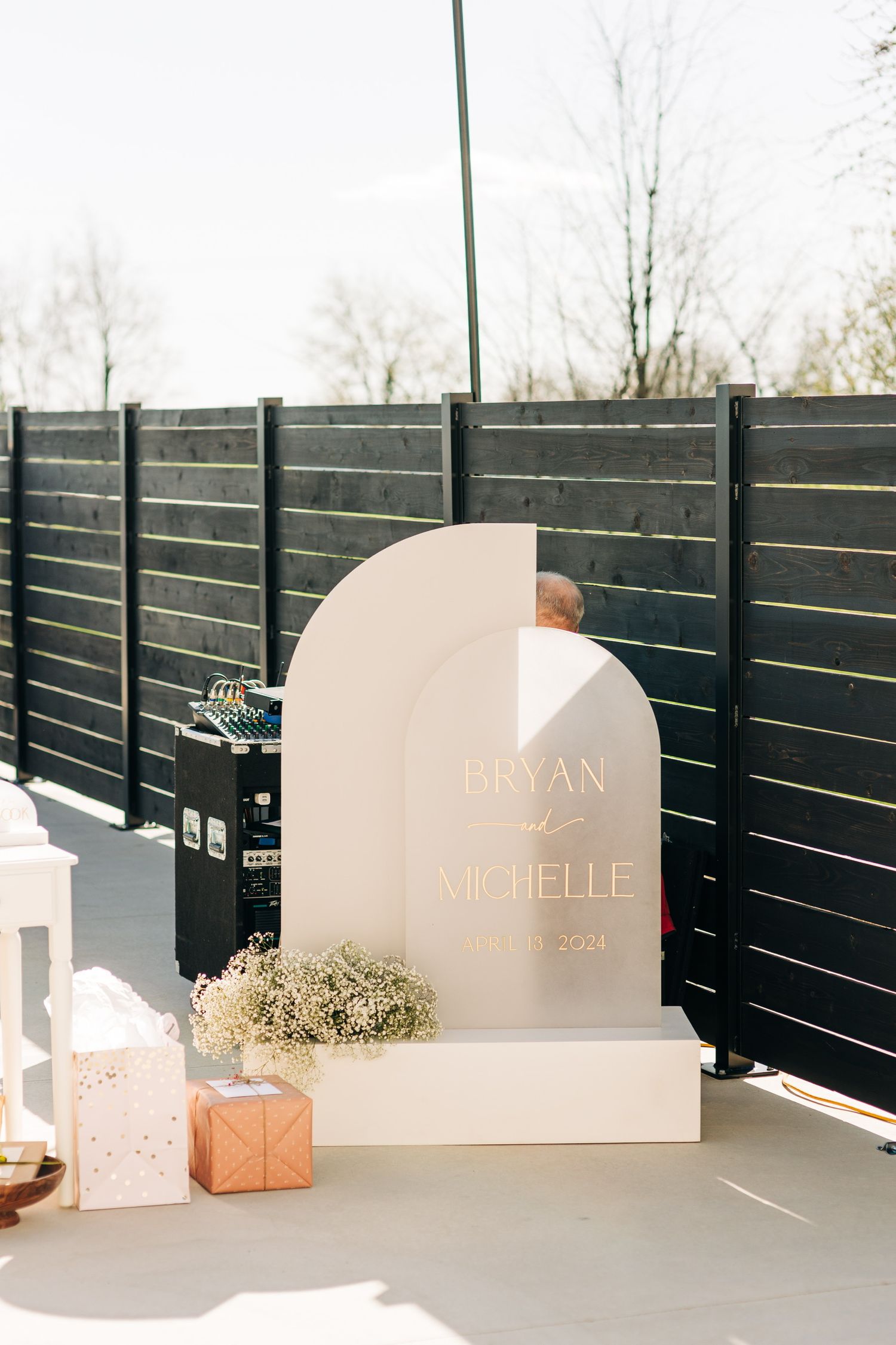 A modern curved wedding welcome sign against a dark wood fence with decorative accessories.