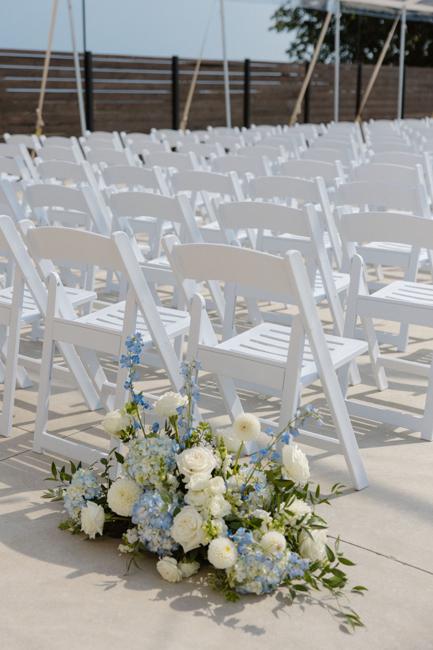 Rows of white folding chairs at an outdoor wedding ceremony with a delicate blue and white floral arrangement on the aisle.