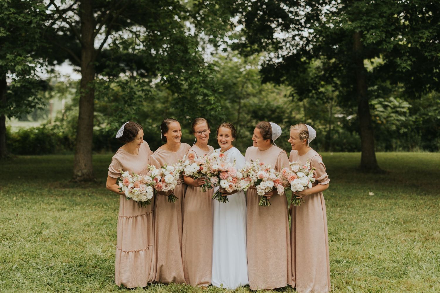 Wedding party members in blush pink dresses pose together outdoors with floral bouquets in a garden setting.