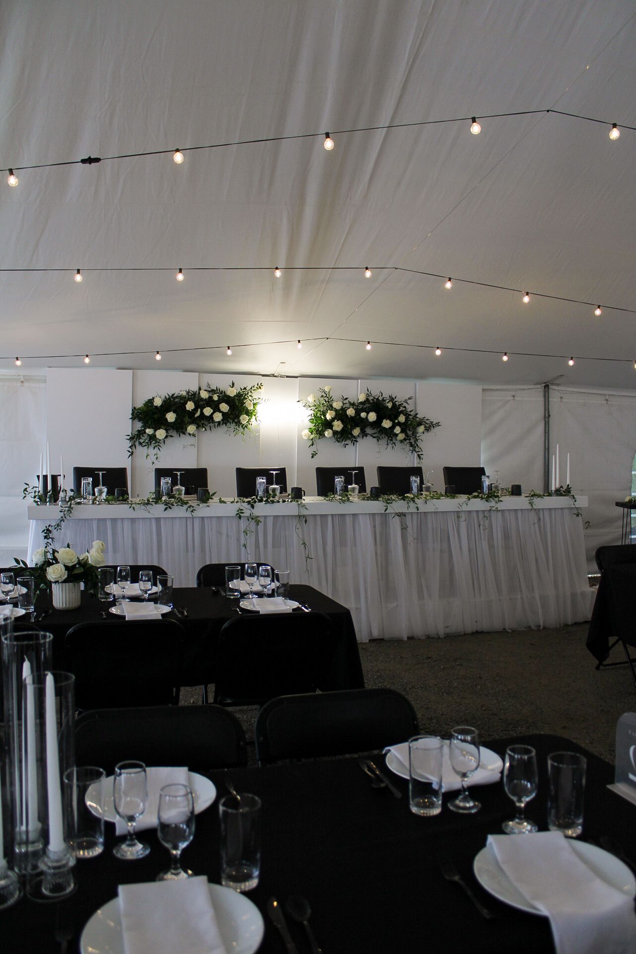 Wedding reception tent with string lights, black tablecloths, and white floral head table decorations for an elegant setup.