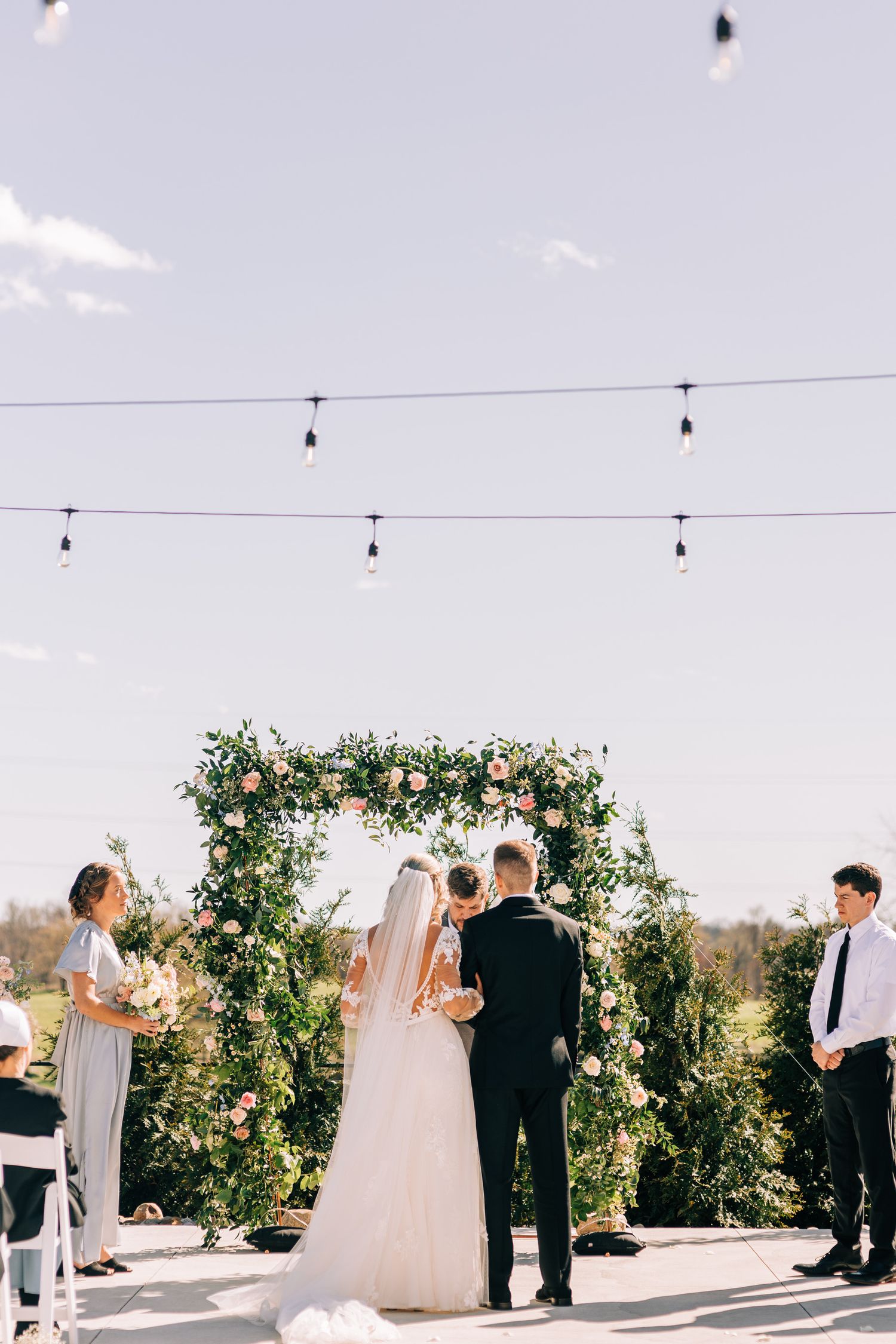 A lush floral wedding arch with string lights overhead during an outdoor ceremony.