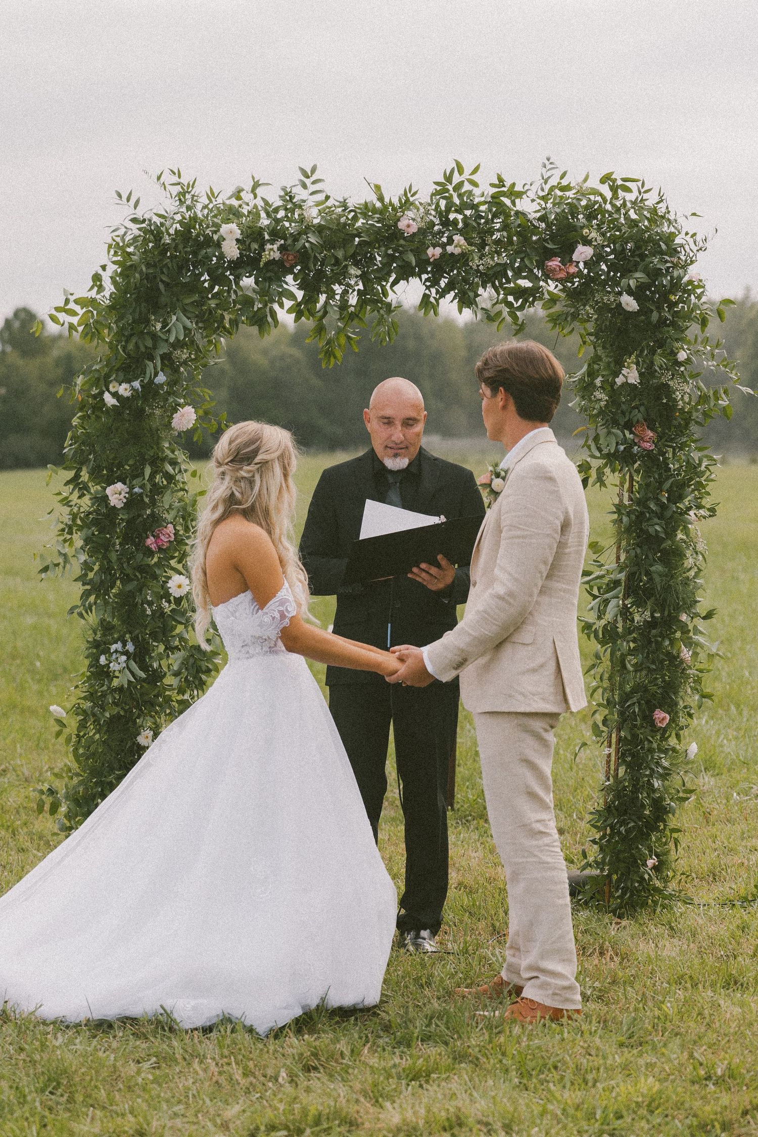 Wedding ceremony beneath floral arch with roses and greenery in countryside setting.