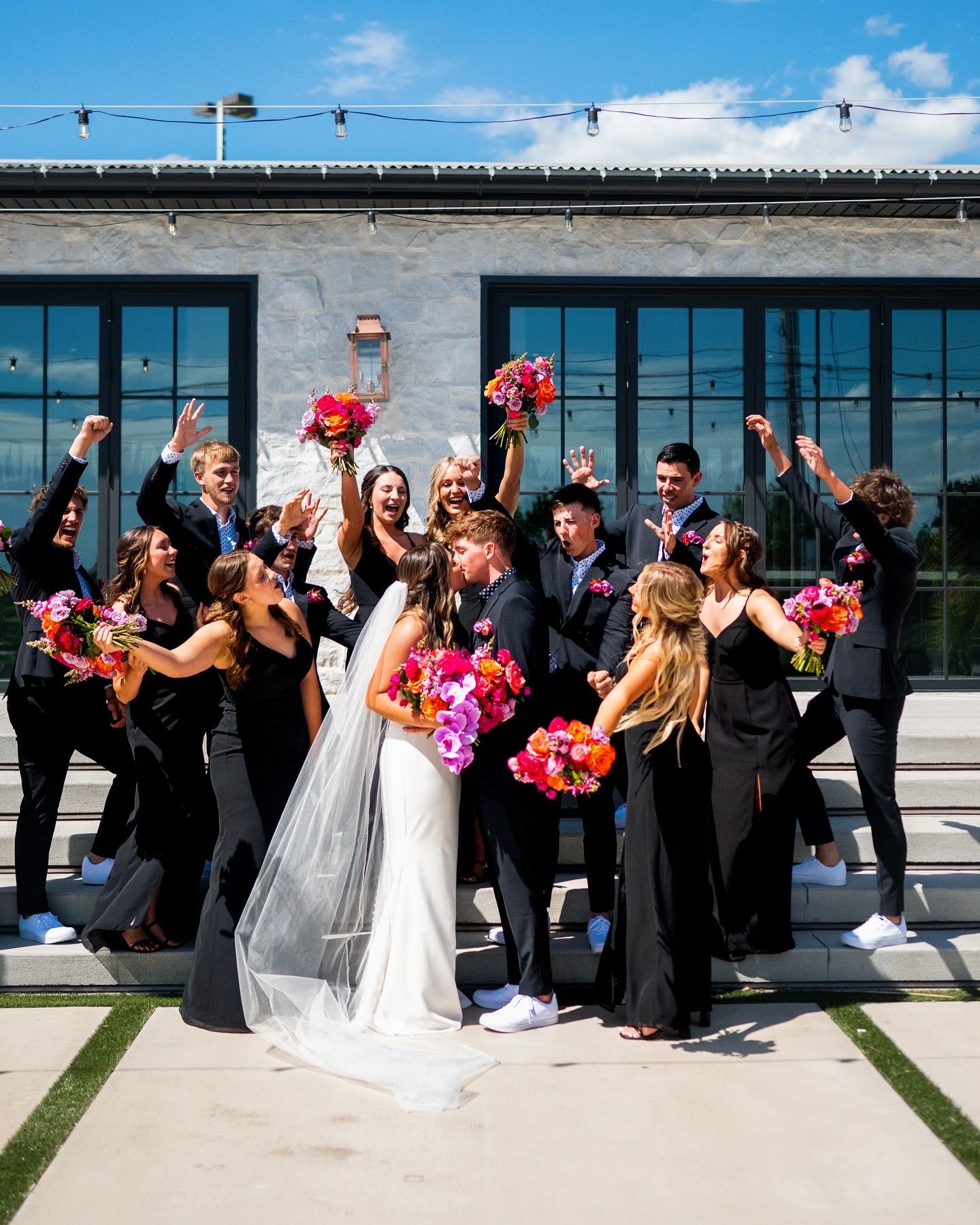 Energetic wedding party celebration photo outside modern venue with bright bouquets.