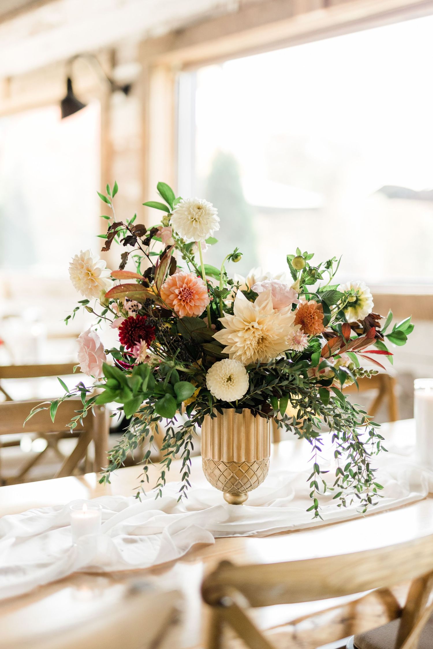 Romantic floral arrangement with dahlias and greenery in gold vase on sunlit table.