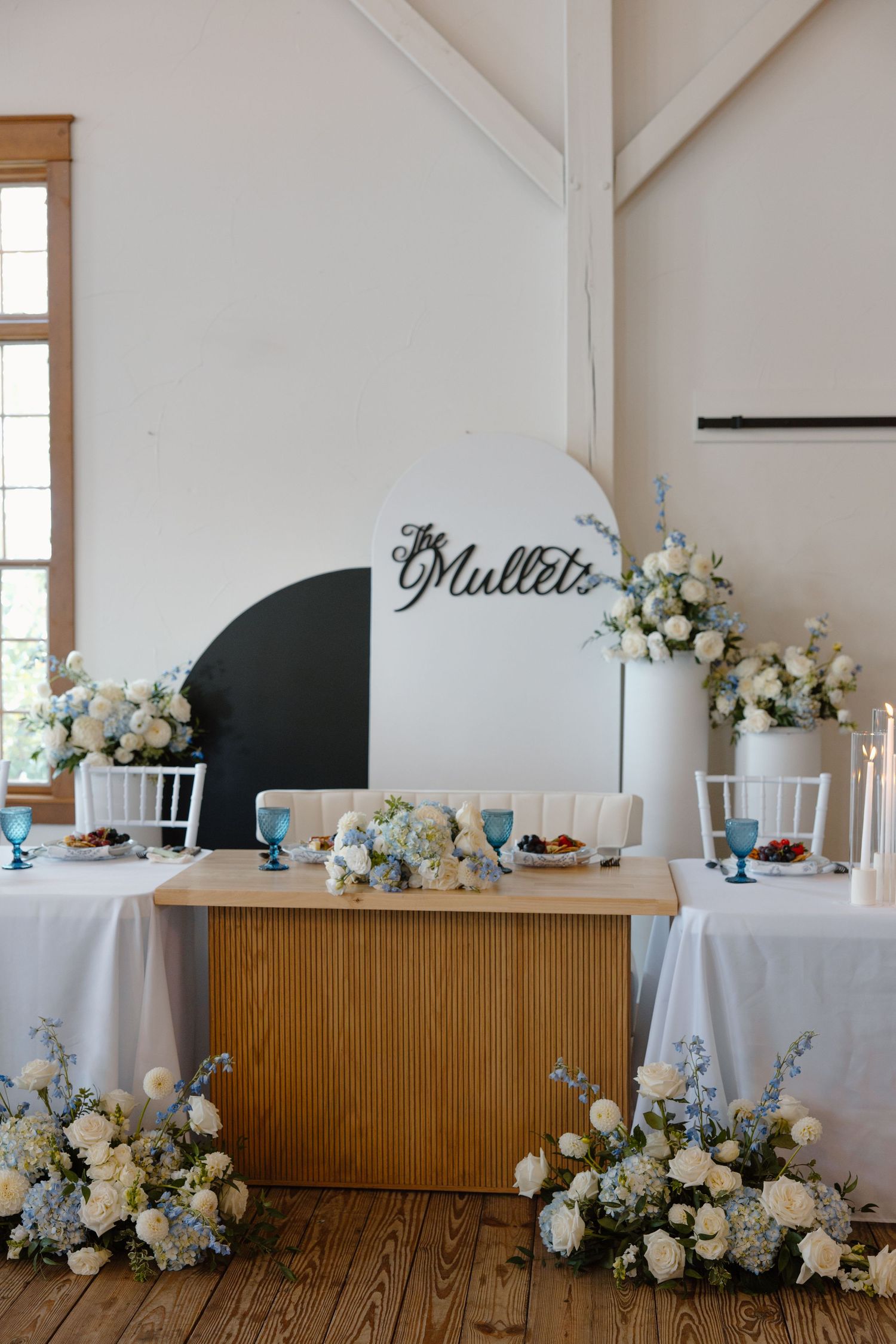 Elegant wedding head table with blue and white floral arrangements against a white wall.
