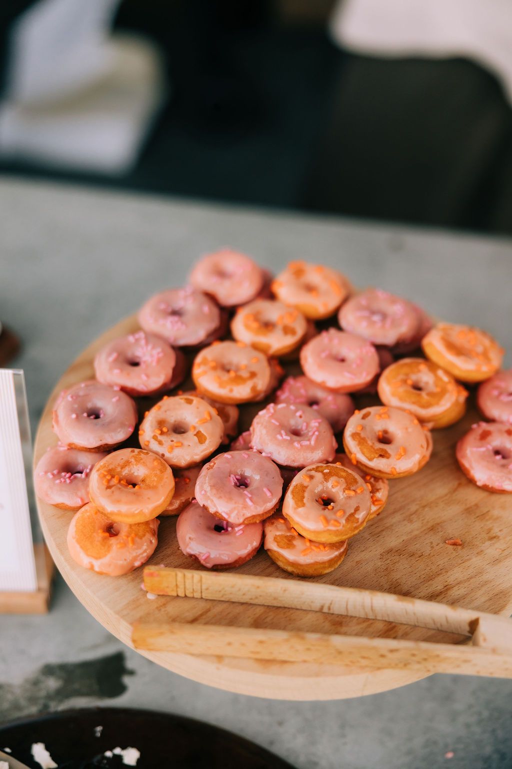 Pink and orange frosted mini donuts arranged on a round wooden serving board with wooden utensils.