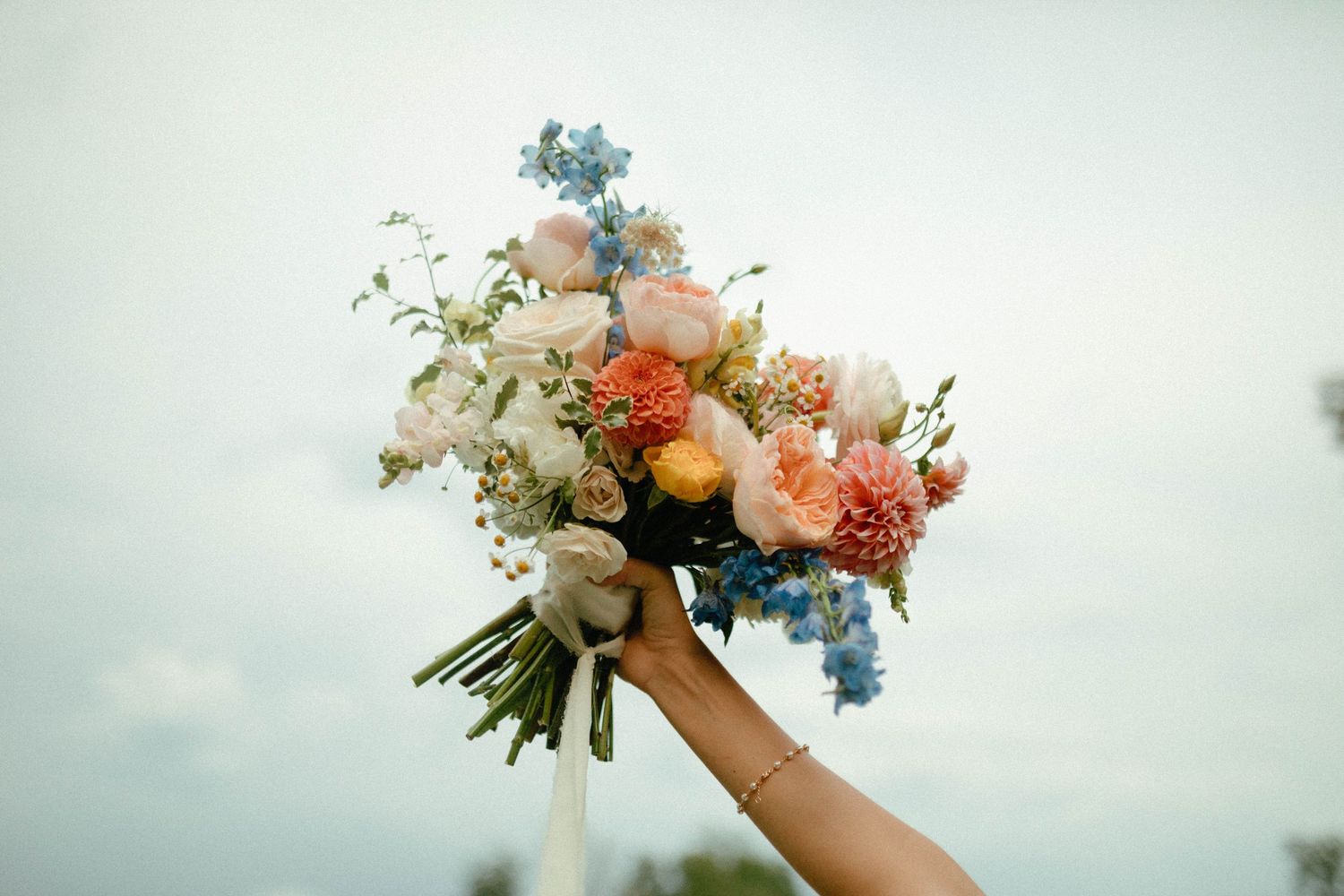 A delicate peach and blue floral bouquet with roses and delphiniums is held up against a cloudy sky.