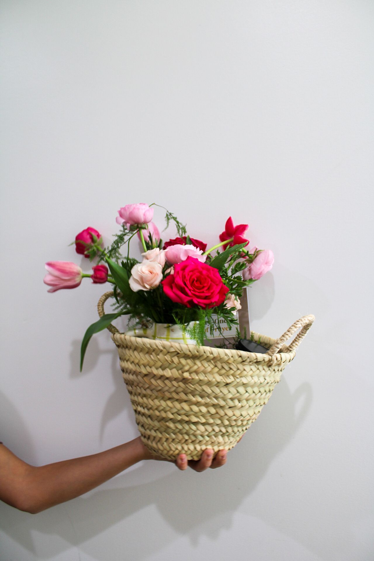 Hand holding a woven straw basket filled with pink and red roses and mixed flowers against a white wall.