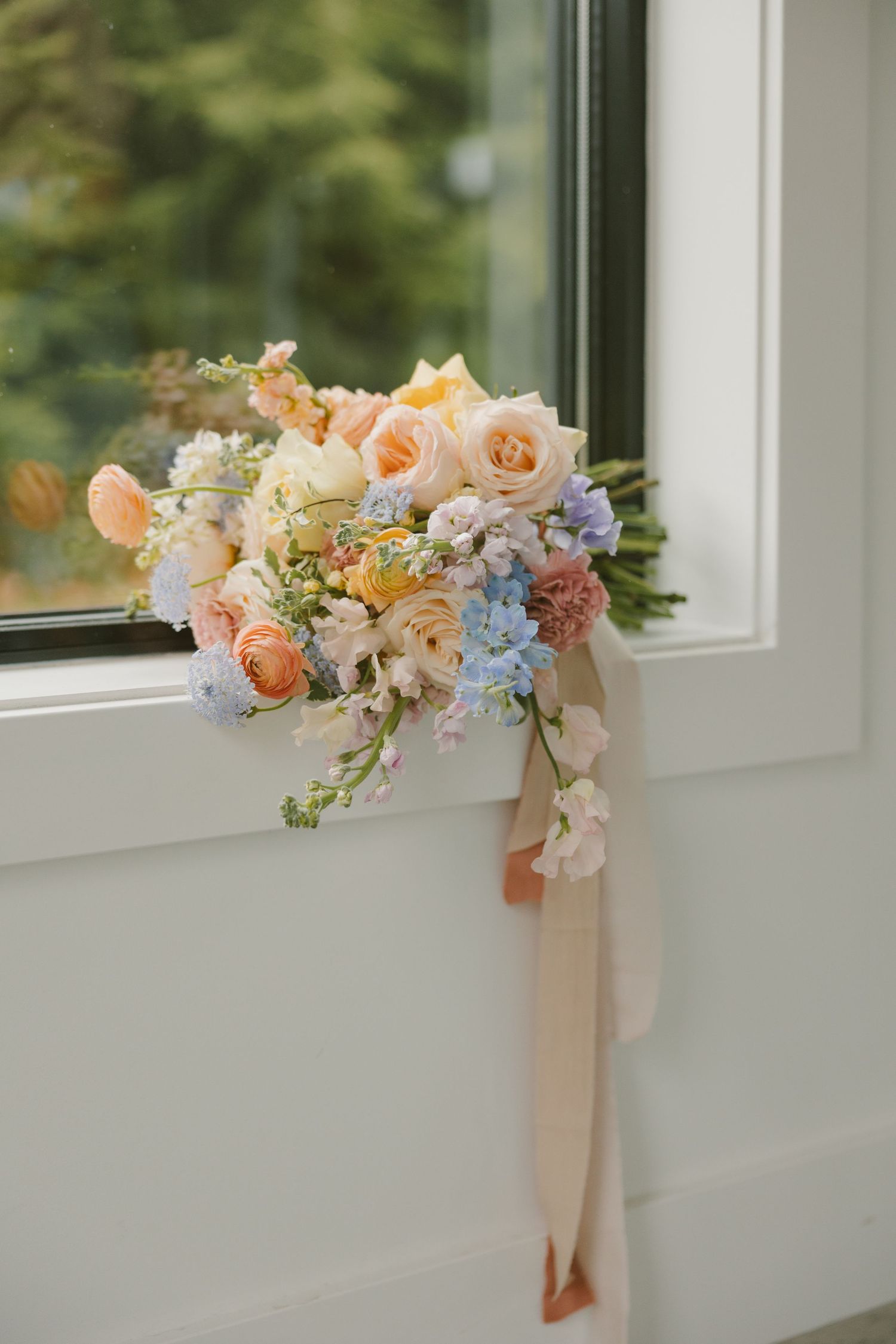 Romantic pastel bouquet with peach roses and blue accents sitting on a white windowsill.