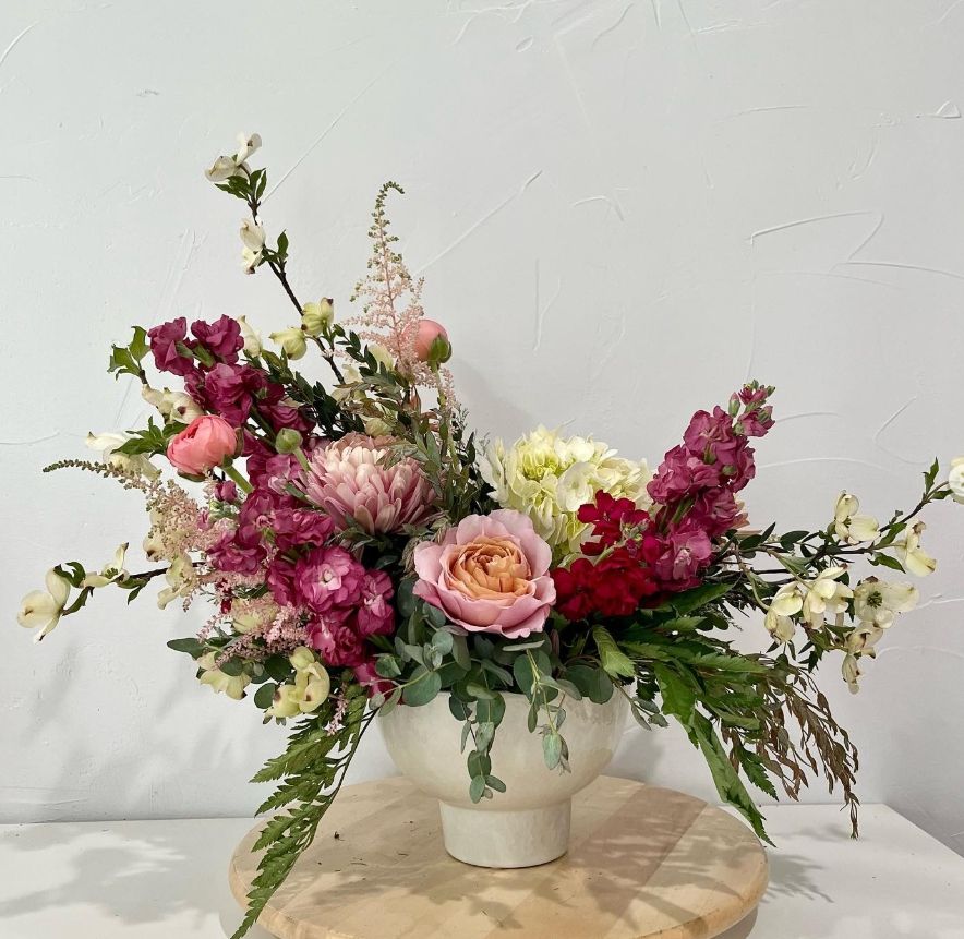 Romantic pink and burgundy flower arrangement with roses and greenery in a white ceramic container.