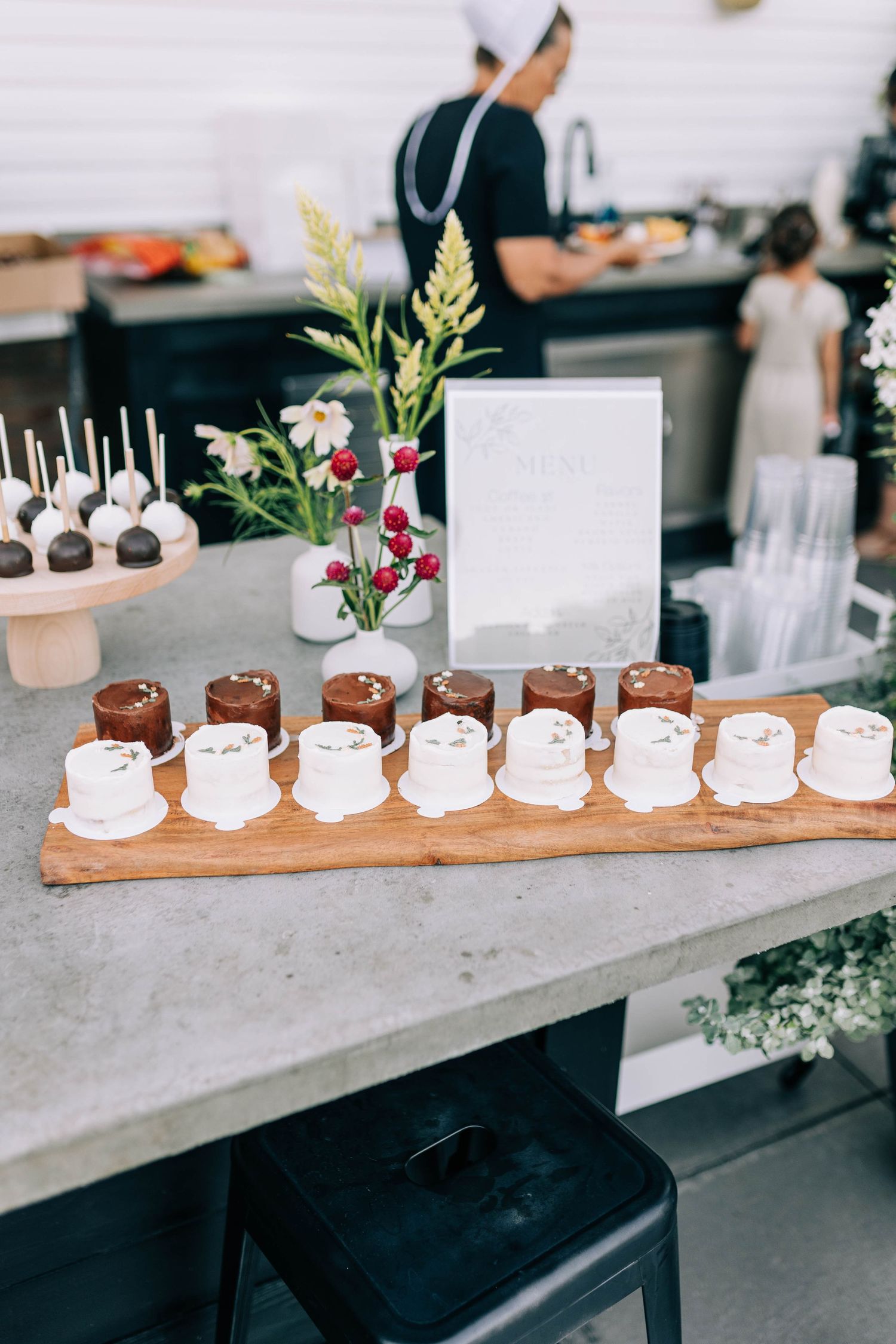 Chocolate cake pops and desserts displayed on wooden board with floral decorations at catered event.