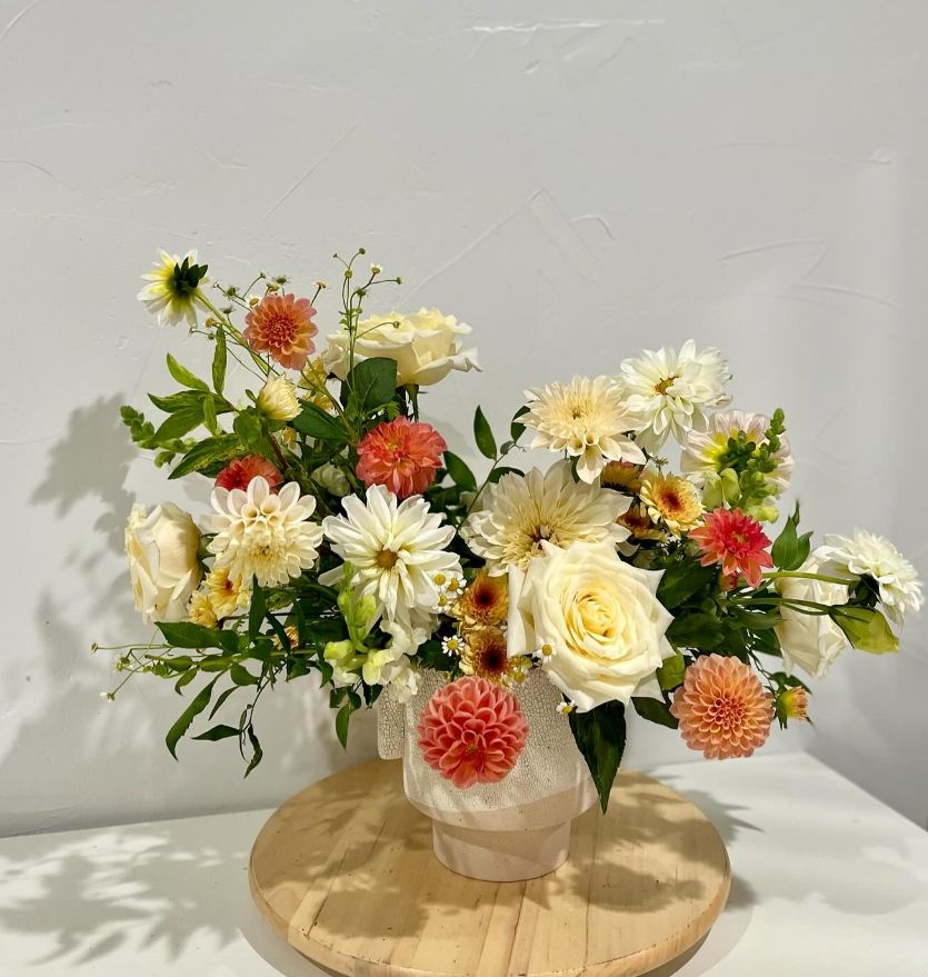 Cheerful arrangement of coral dahlias, cream roses, and daisies in a frosted vase on wooden platform.