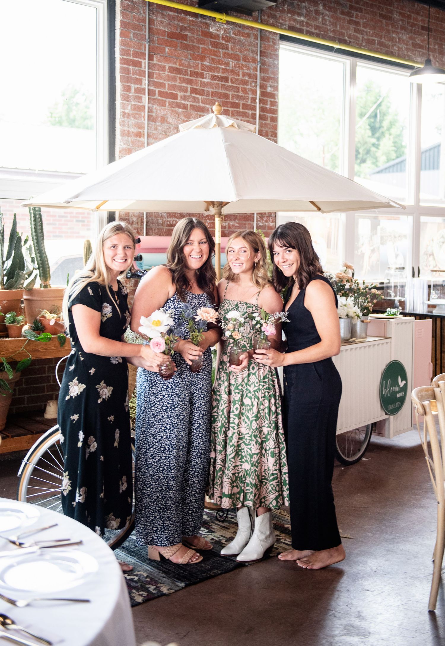 Group gathering under white umbrella with floral arrangements and vintage bicycle in industrial space.