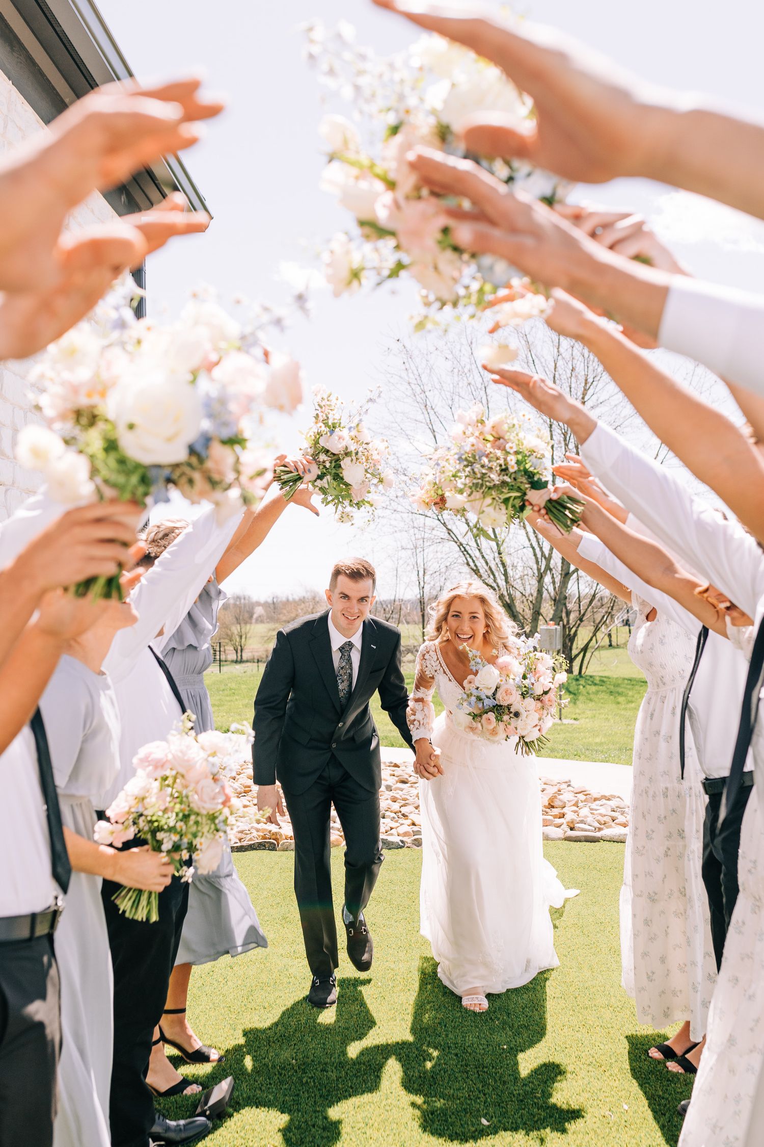 Wedding party creates flower petal tunnel arch for newlyweds exiting ceremony on green lawn.