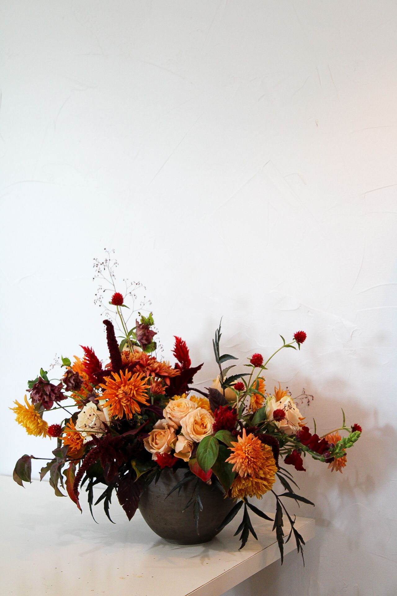 Vibrant autumn floral arrangement with orange dahlias and roses in a dark round vessel against white wall.