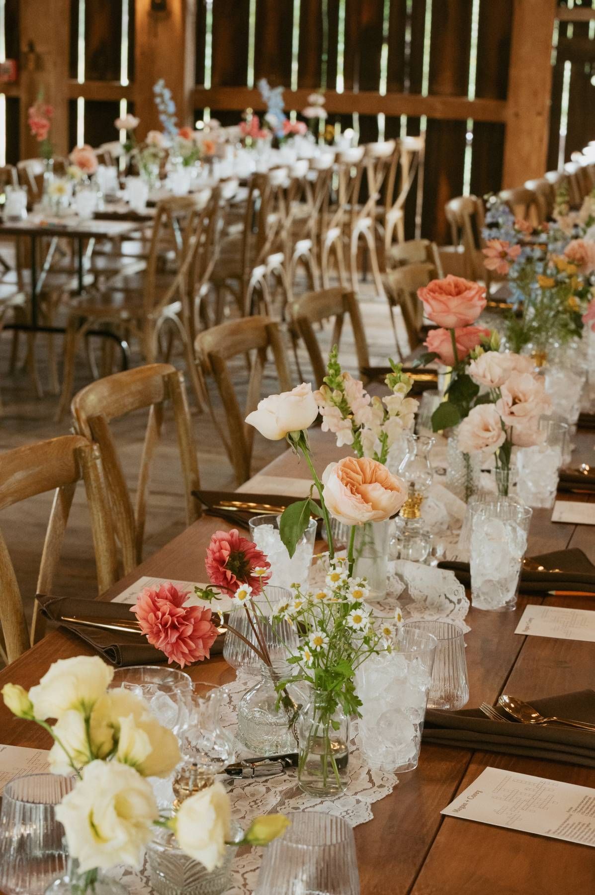 Elegant wedding reception table setting with pink and white flowers, crystal glasses, and rustic wooden chairs.