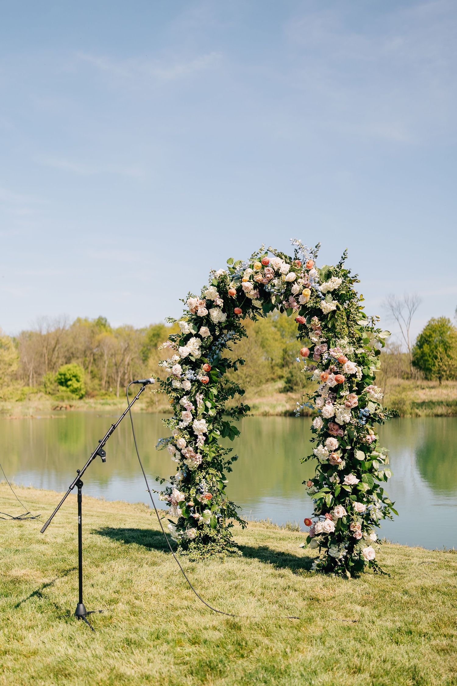 A floral wedding arch with pink and white flowers stands on a grassy hill overlooking a serene lake on a sunny day.