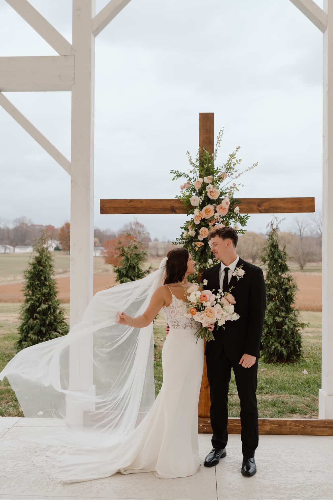 Romantic wedding scene at outdoor chapel with flowing white dress and wooden cross against rural landscape.