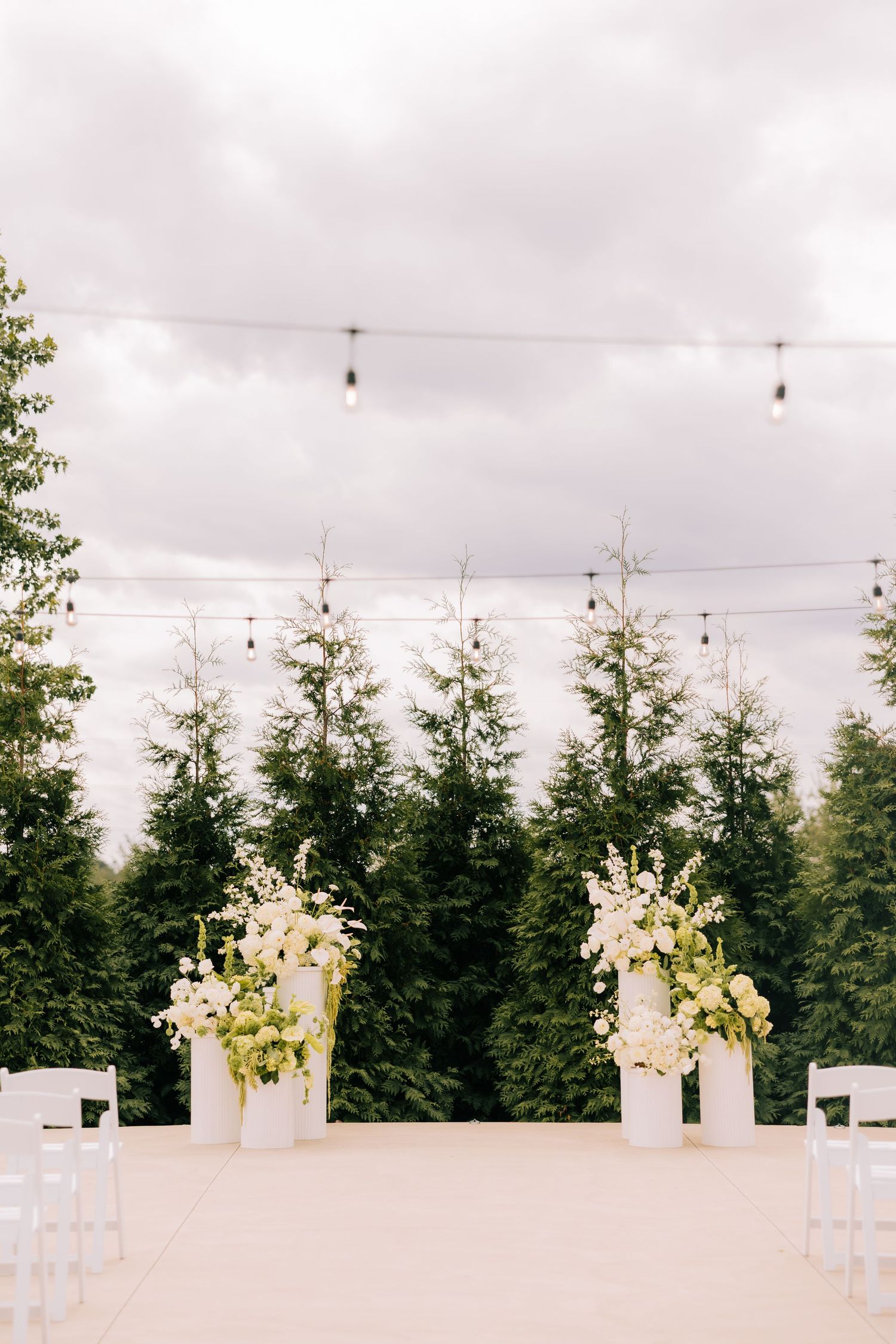White floral arrangements in tall vases line an outdoor wedding ceremony aisle with evergreen trees in the background.