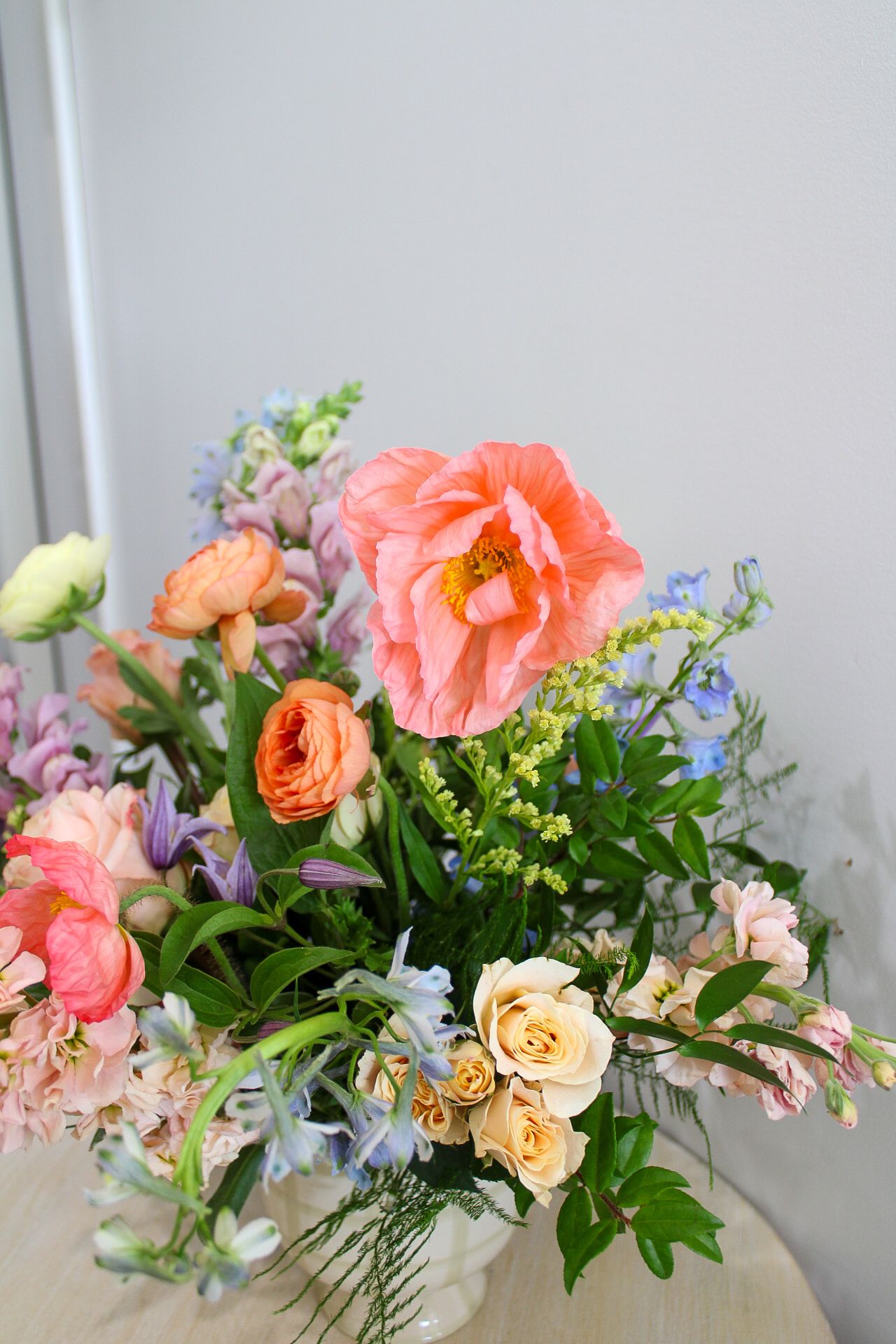 Vibrant spring floral arrangement with pink peony, coral ranunculus, cream roses, and blue flowers in a white vase.