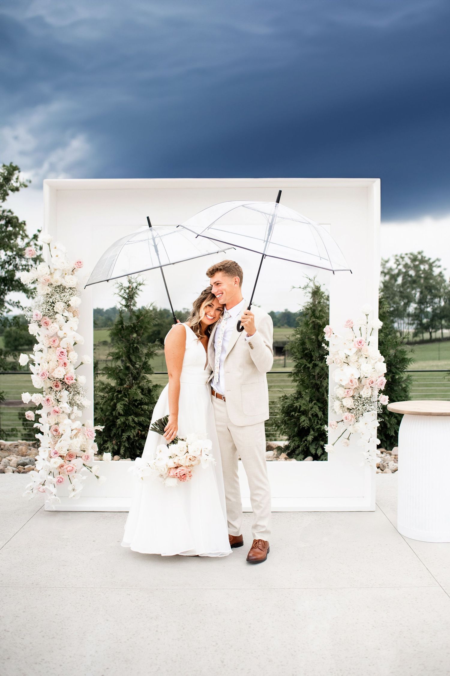 Wedding couple poses with clear umbrella against stormy sky and white floral arch.