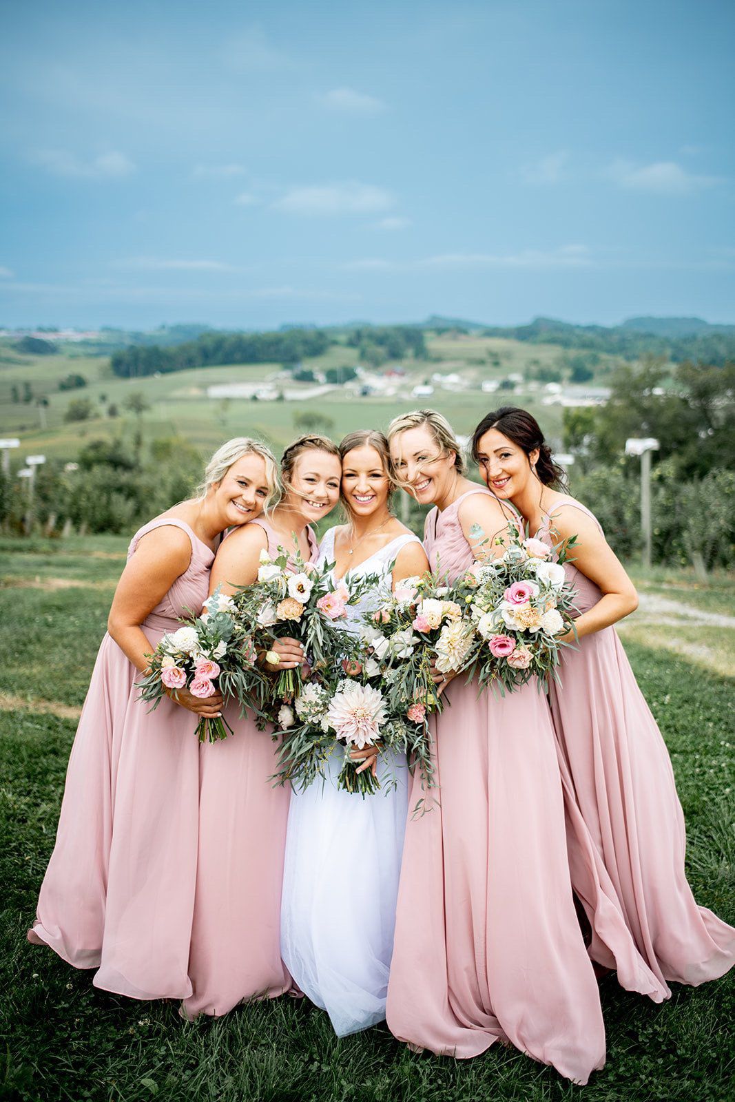 Bridesmaids in flowing pink gowns pose with bride on scenic hilltop with floral bouquets.