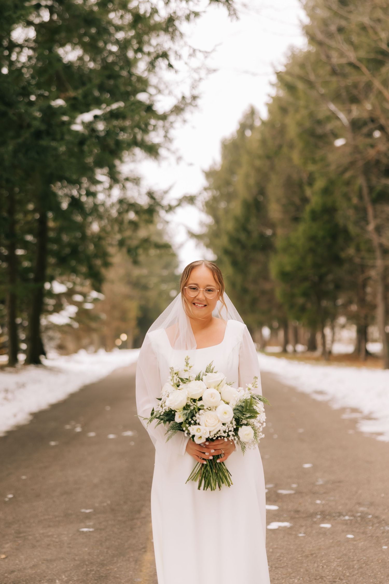 A bride in a flowing white dress holds a winter bouquet while walking along a snowy forest path.