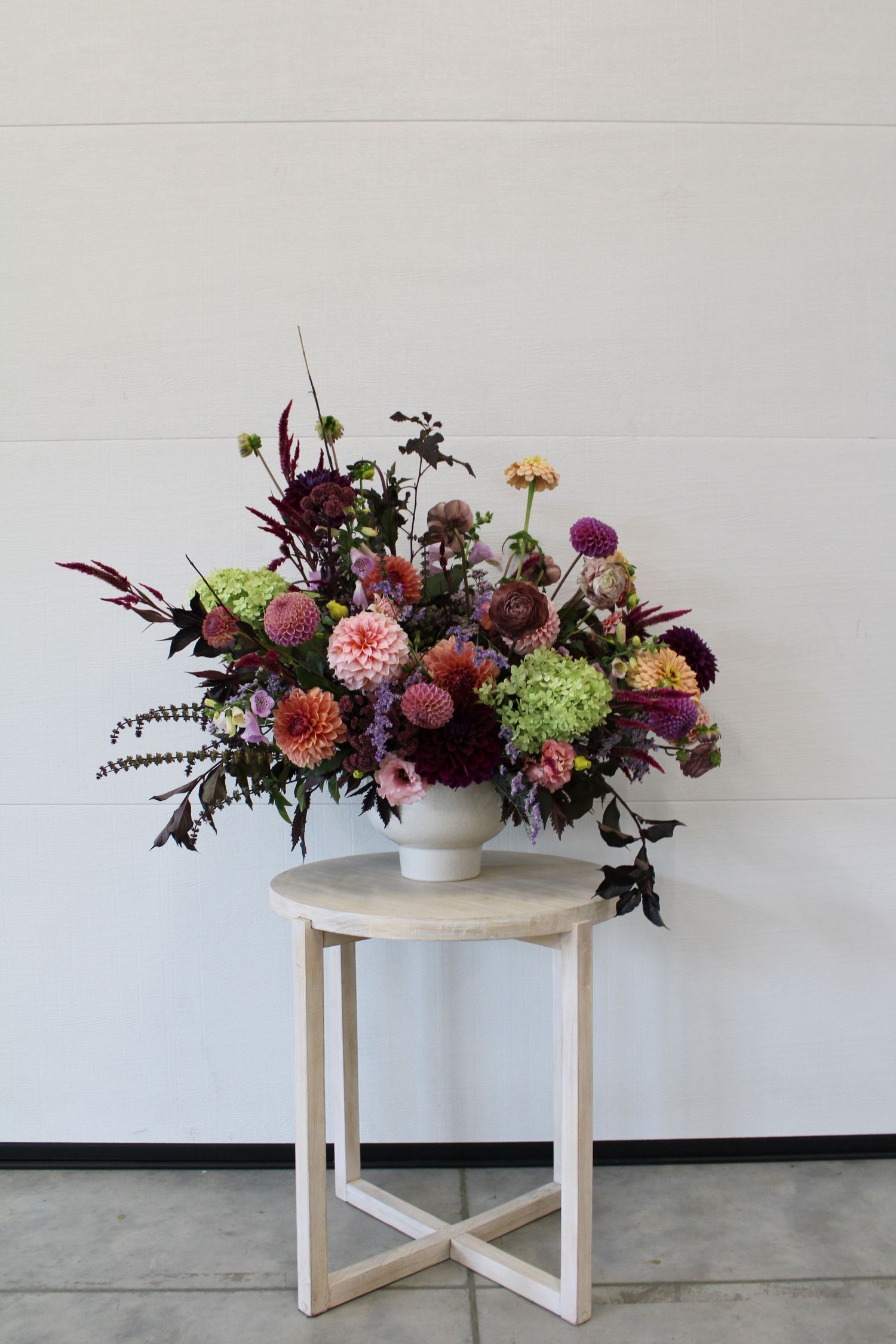 A modern floral arrangement with dahlias and greenery sits atop a white round side table against a light gray wall.