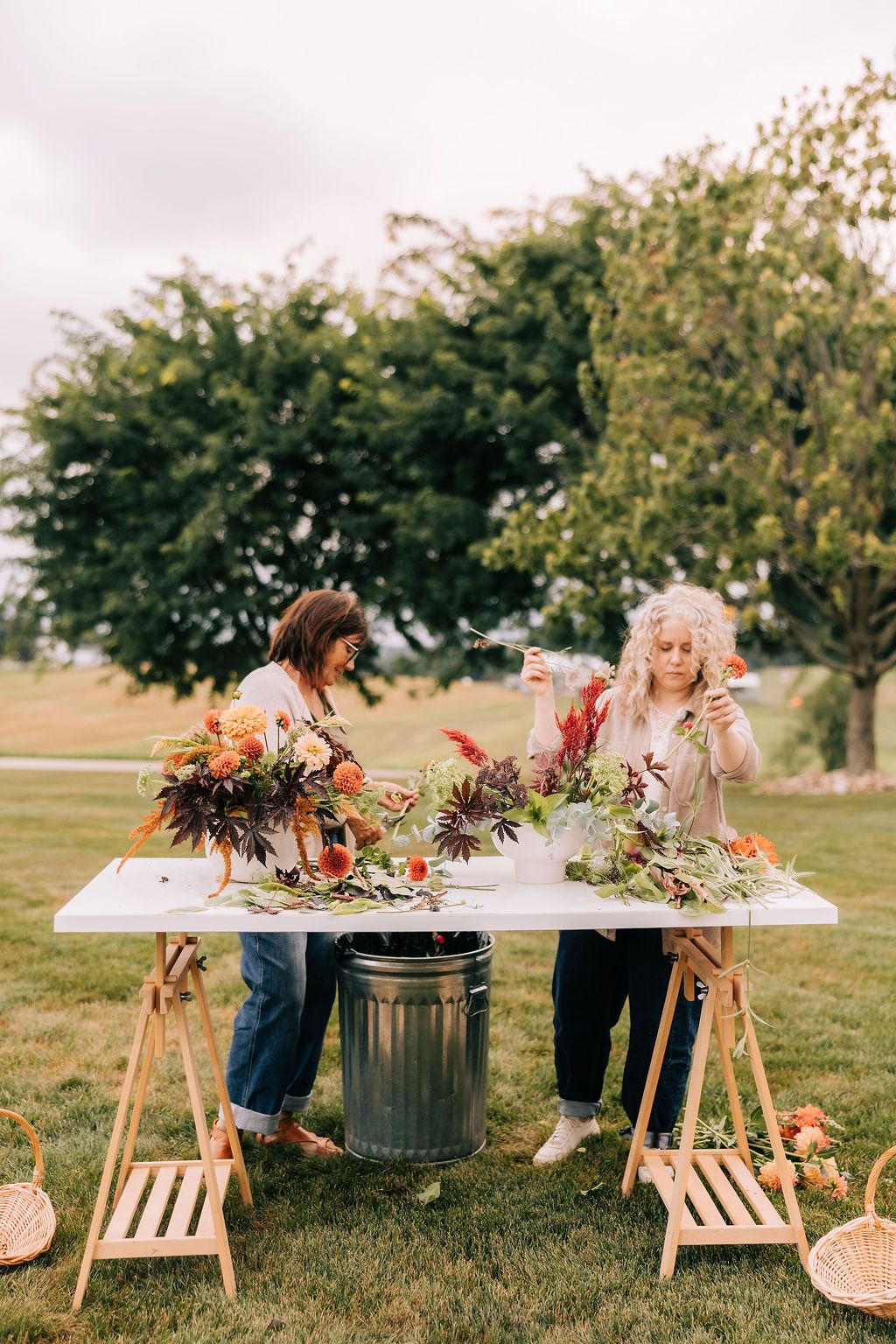 People gather around wooden tables decorated with autumn floral arrangements and greenery at an outdoor event.