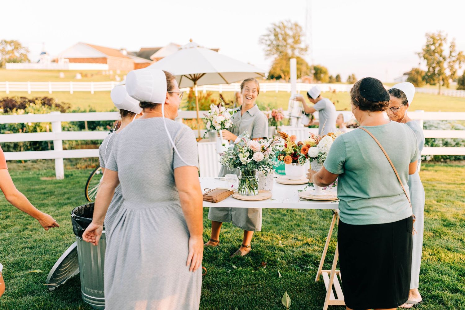 People working at tables outdoors arranging flowers with white fencing and trees in the background.