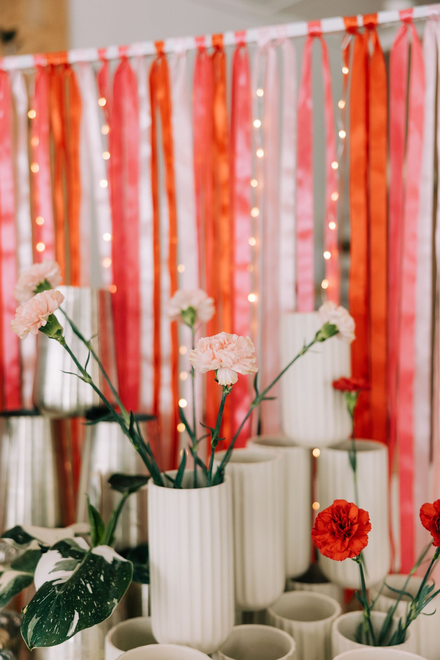 White ceramic vases with carnations against coral and pink ribbon backdrop with twinkle lights.