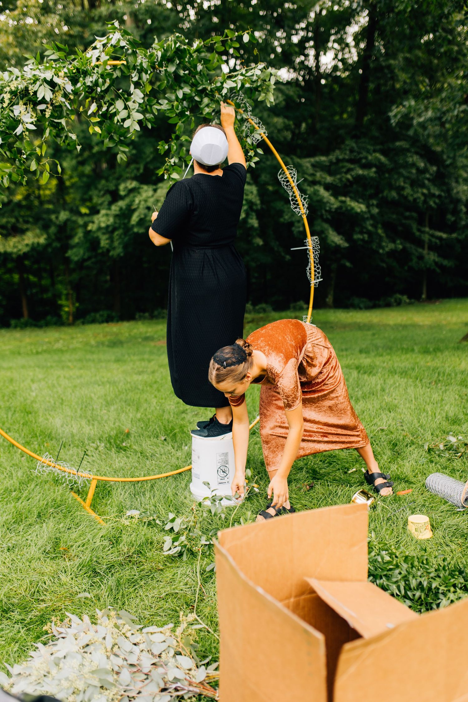 Floral designer in black dress standing on white bucket while arranging outdoor garden display.