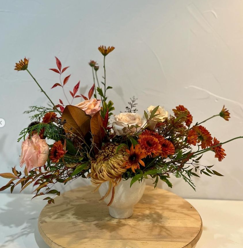 Autumnal floral arrangement with roses and wildflowers in cream ceramic vase on wooden stand.