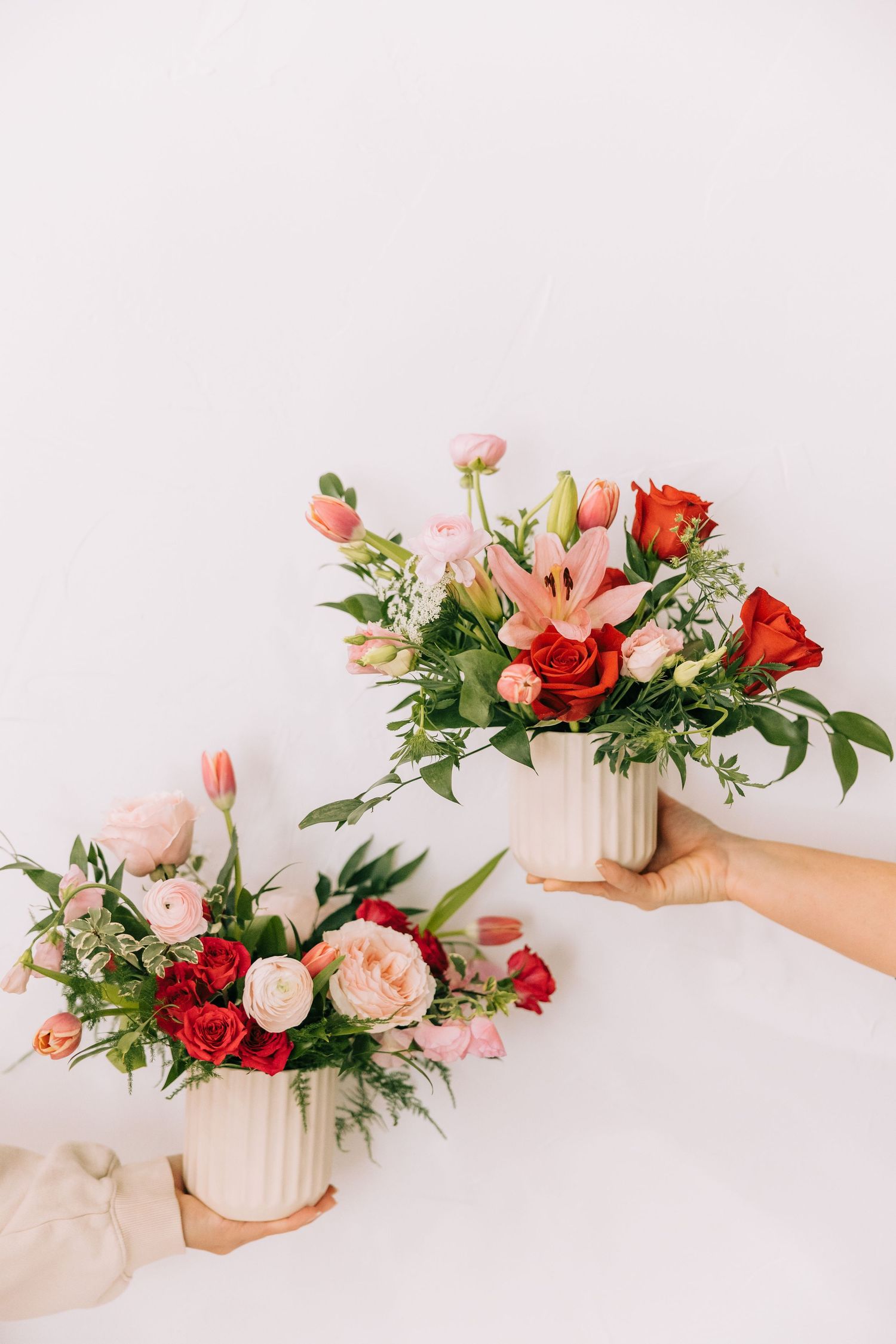Two hands holding matching floral arrangements with pink and red roses in white vases.