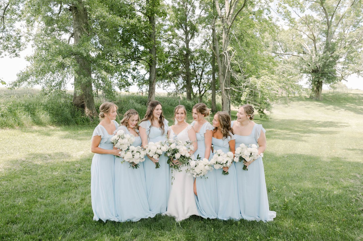 Bridesmaids in pale blue dresses gather for outdoor photo holding matching white bouquets.