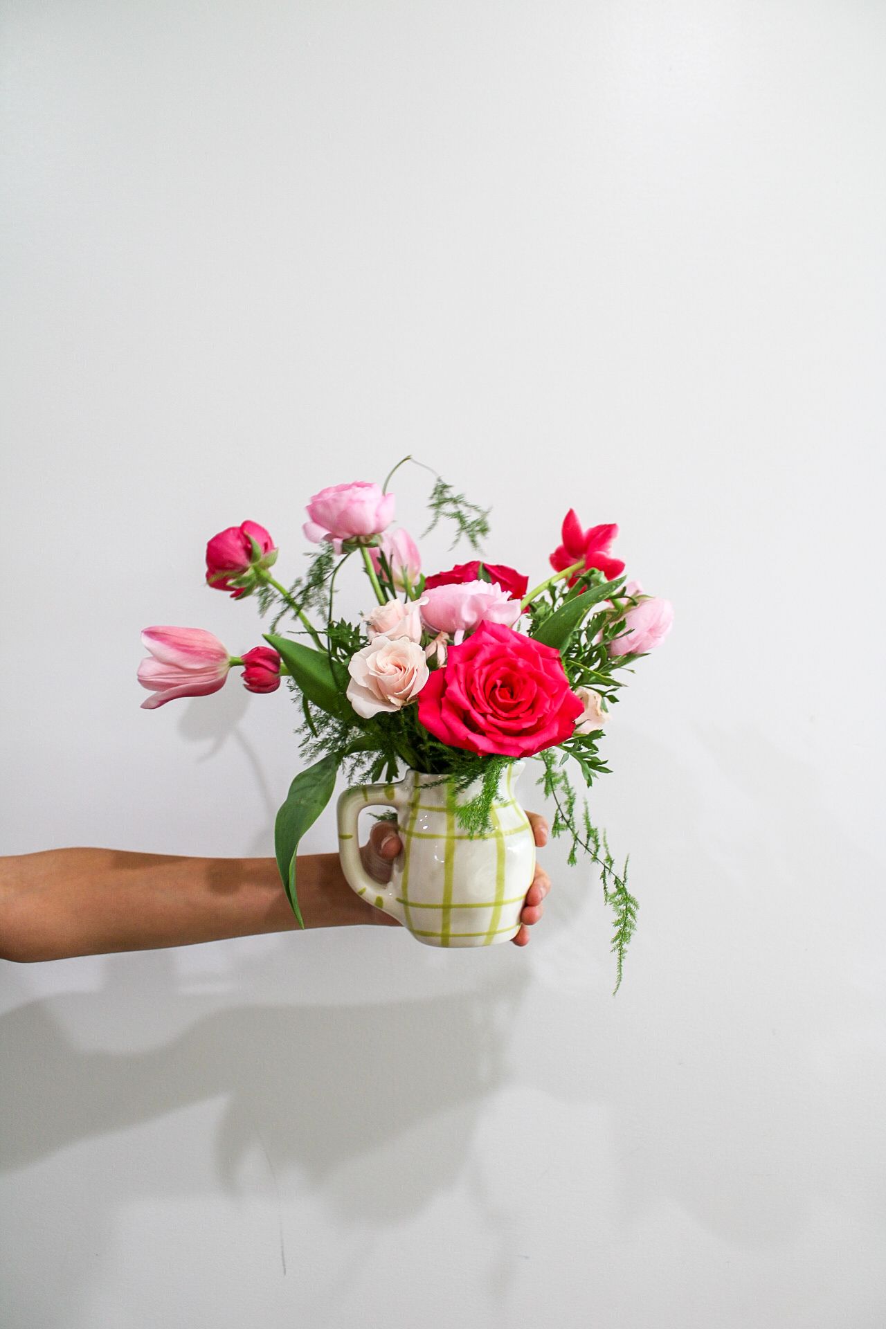 A hand holds a green checkered mug vase filled with pink and red roses, tulips, and ranunculus against a white background.