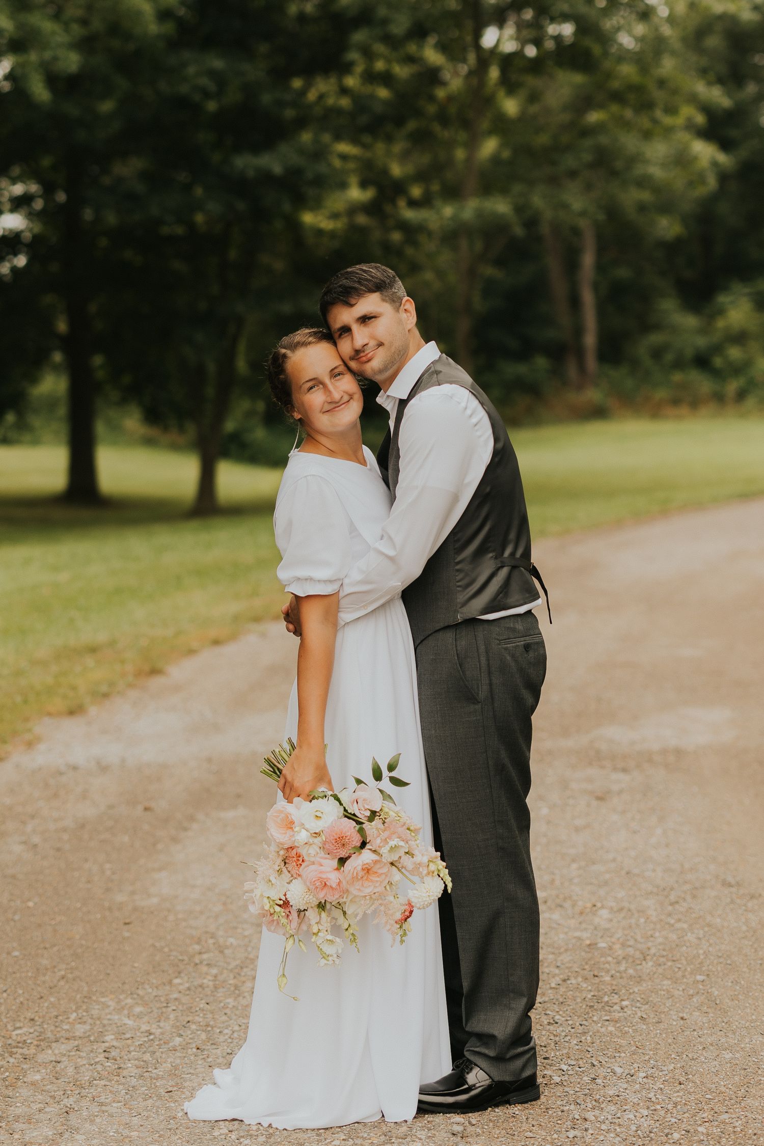 A romantic wedding couple embraces on a tree-lined gravel path while holding a pink and white floral bouquet.