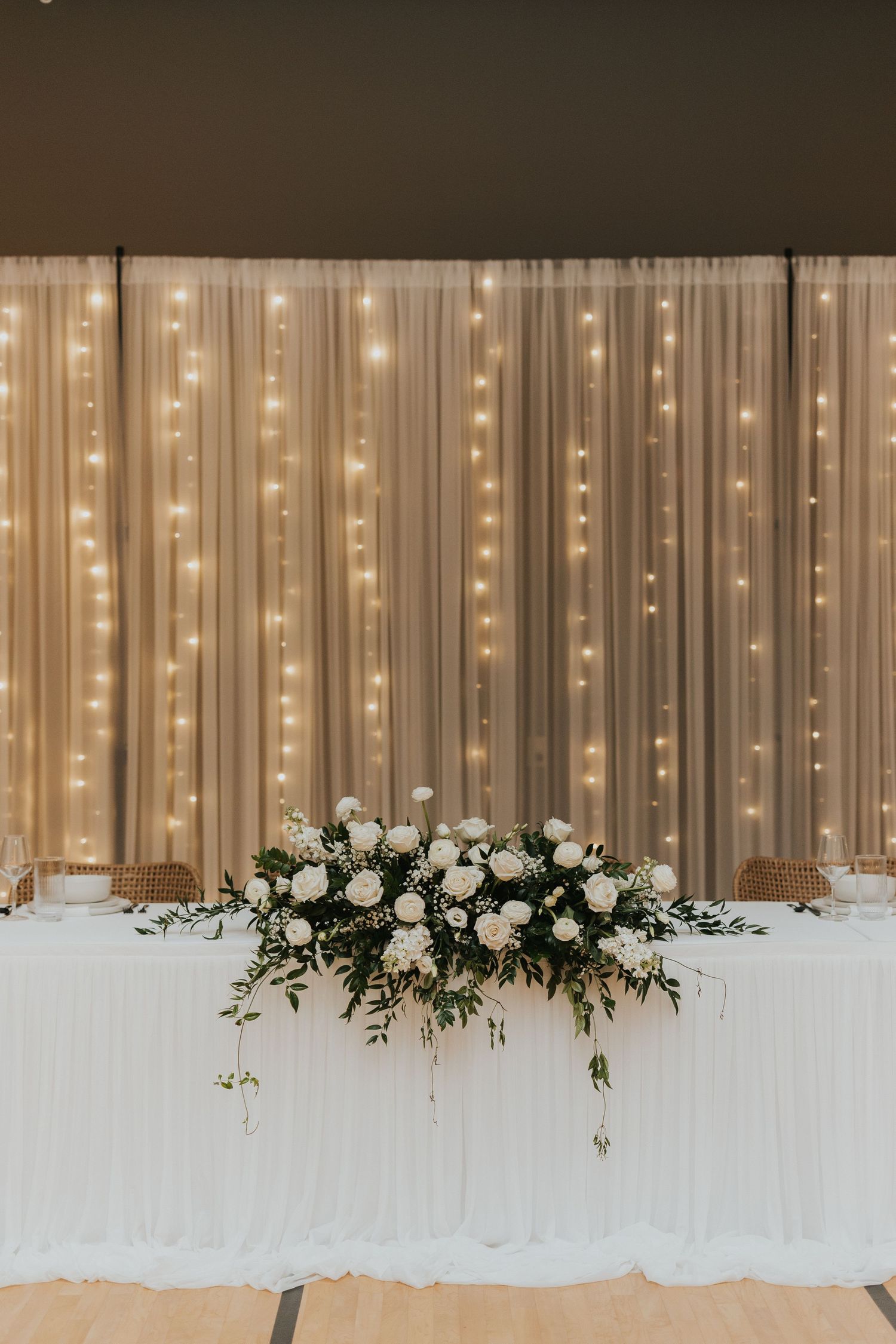 Elegant wedding head table with white floral centerpiece and romantic fairy light backdrop curtain.