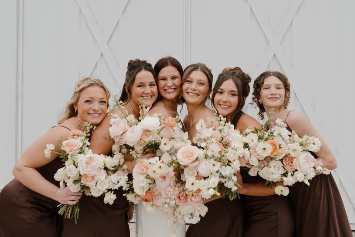 Wedding party poses with peach and cream floral bouquets while wearing brown dresses and white gown.