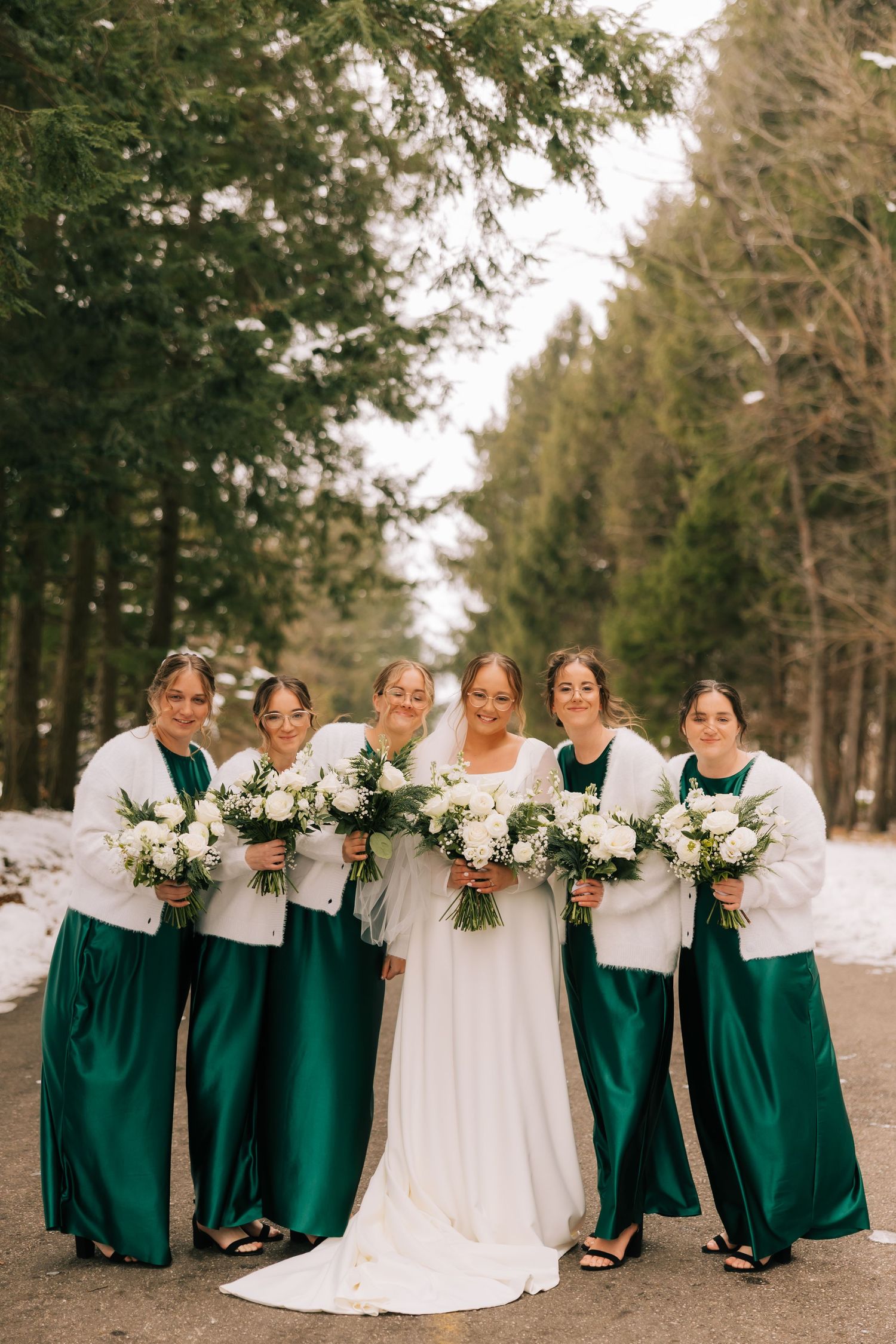A wedding party wearing emerald green dresses and white shawls poses together in a snowy forest setting.
