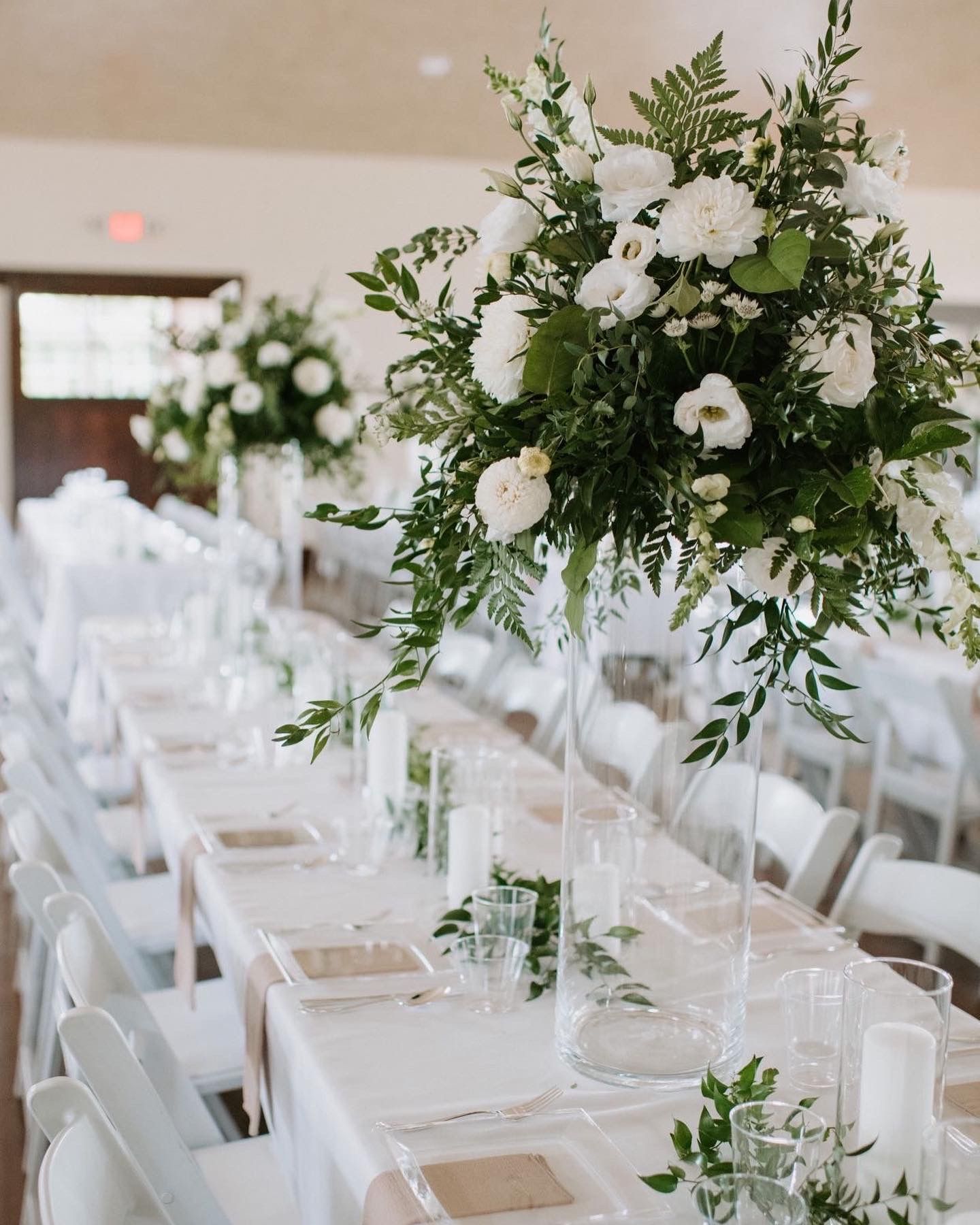 Elegant white wedding reception tables with tall floral centerpieces and white chairs.