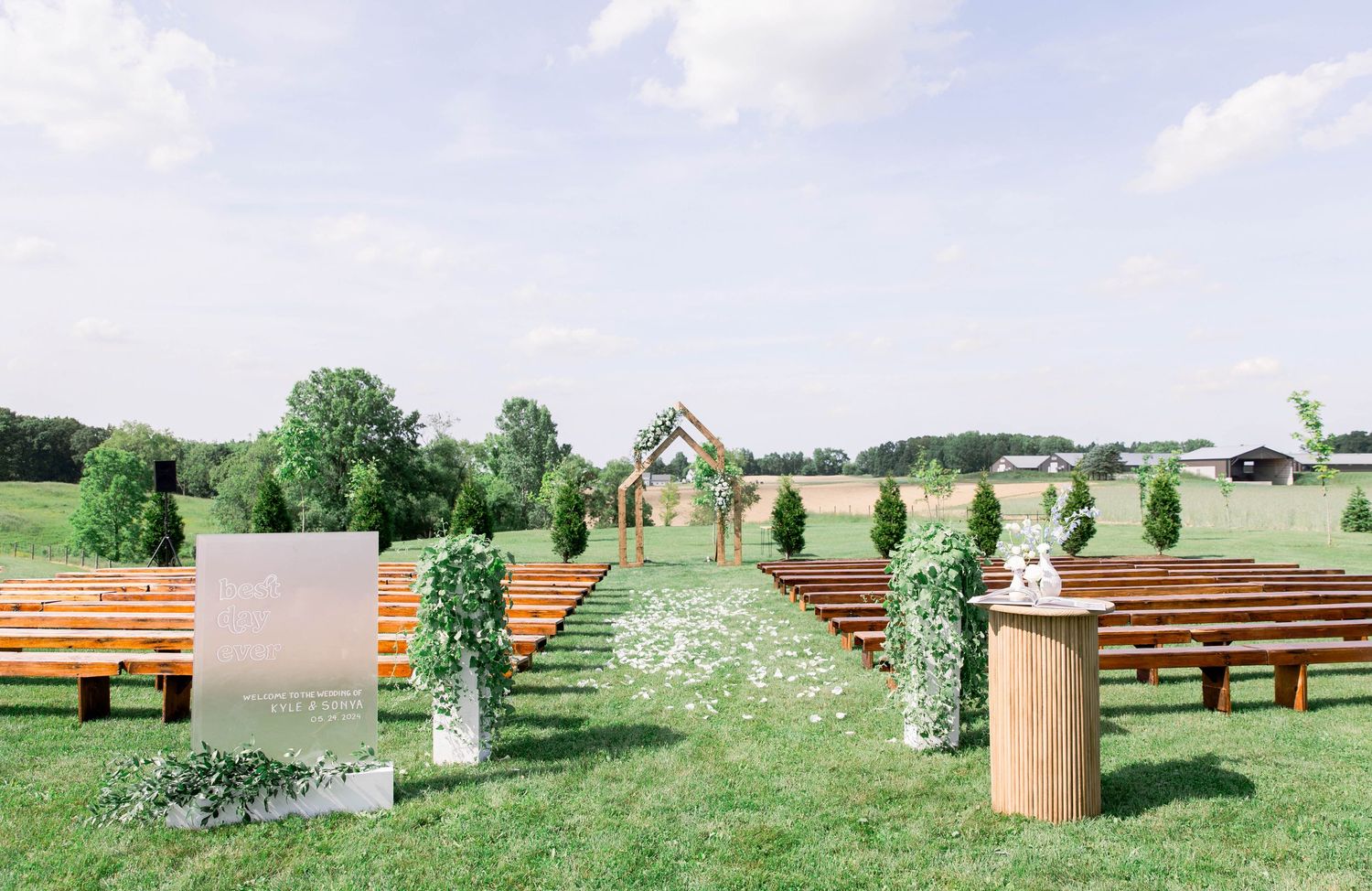 Rustic outdoor wedding venue with wooden benches and simple wooden arch decorated with greenery.